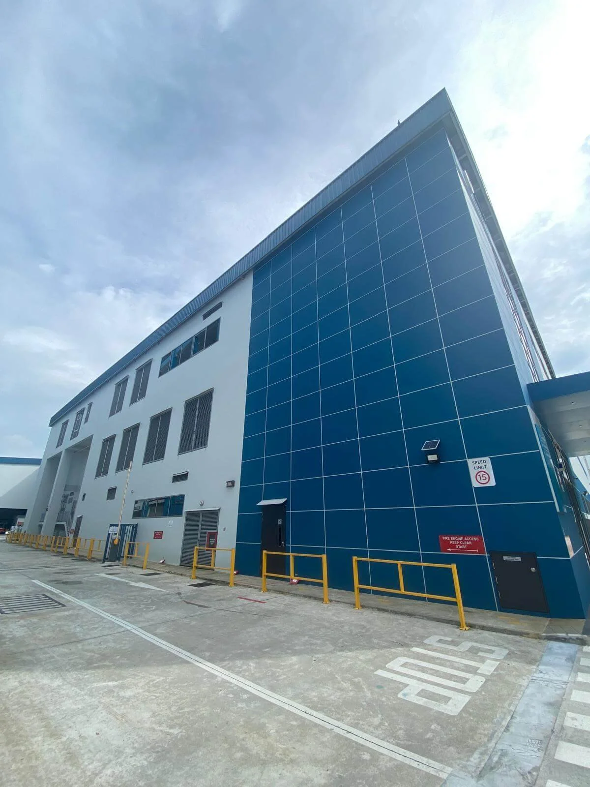 Modern multi-story building with white and blue exterior, parking area with yellow safety barriers, and a speed limit sign of 15 mph.