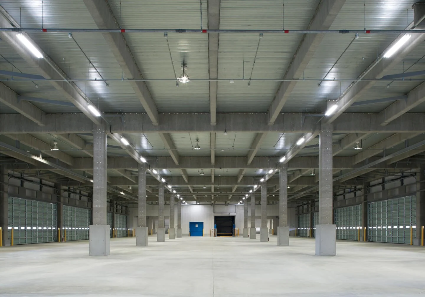 Empty warehouse with concrete floor, metal support beams, overhead lighting, and large garage doors.