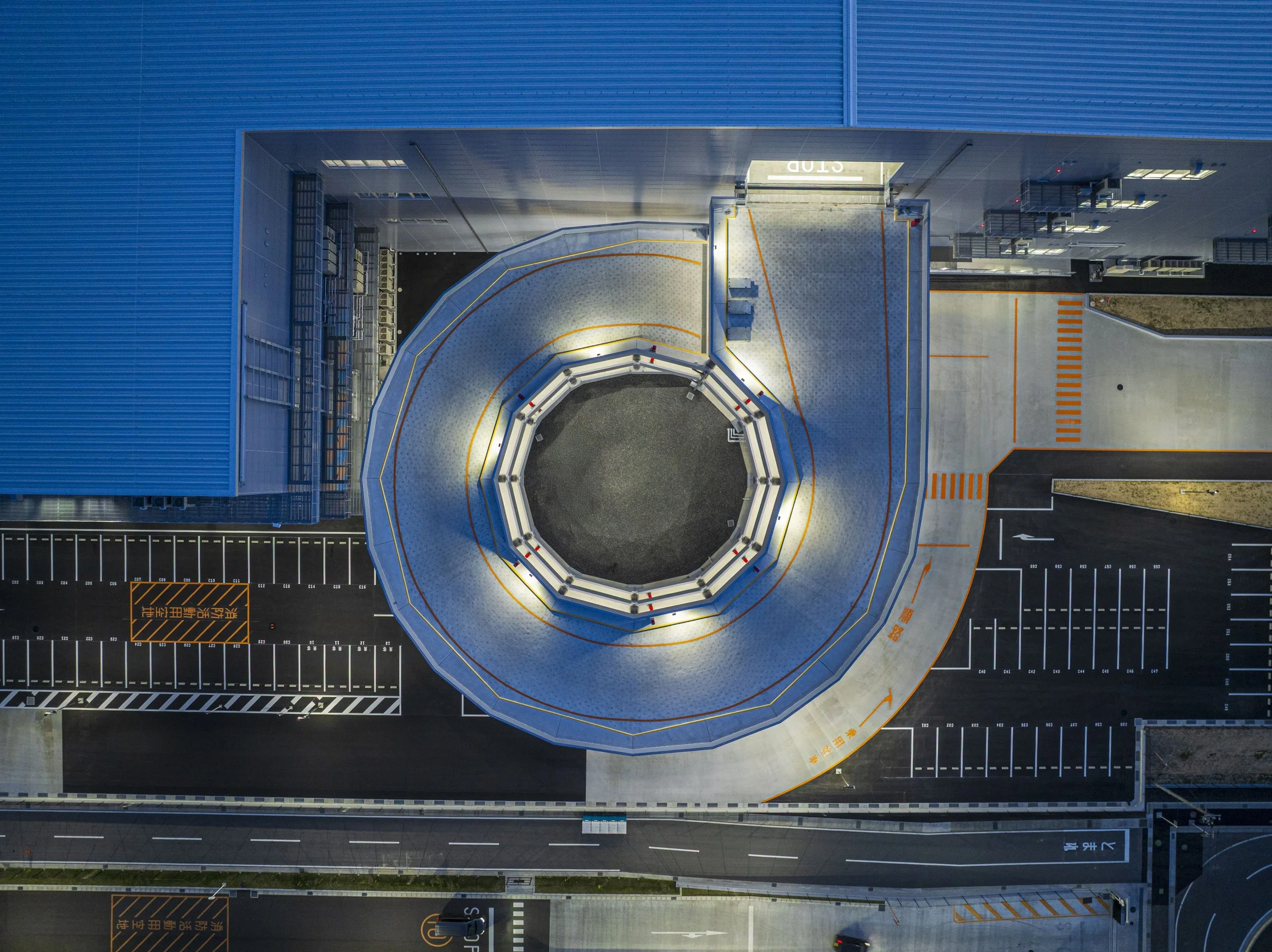 Top-down view of a modern building's rooftop with a circular metallic structure, parking spaces, and surrounding streets.