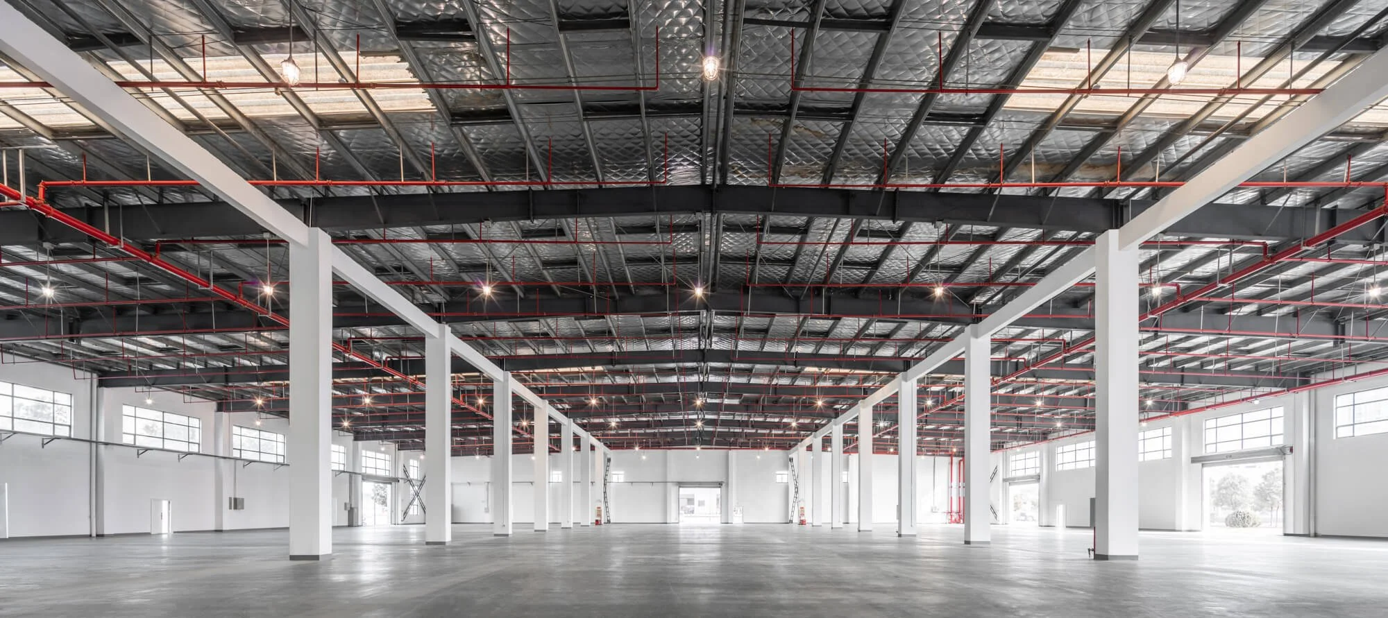 Empty industrial warehouse interior with white columns, large windows, polished concrete floor, and exposed black and red ceiling framework.