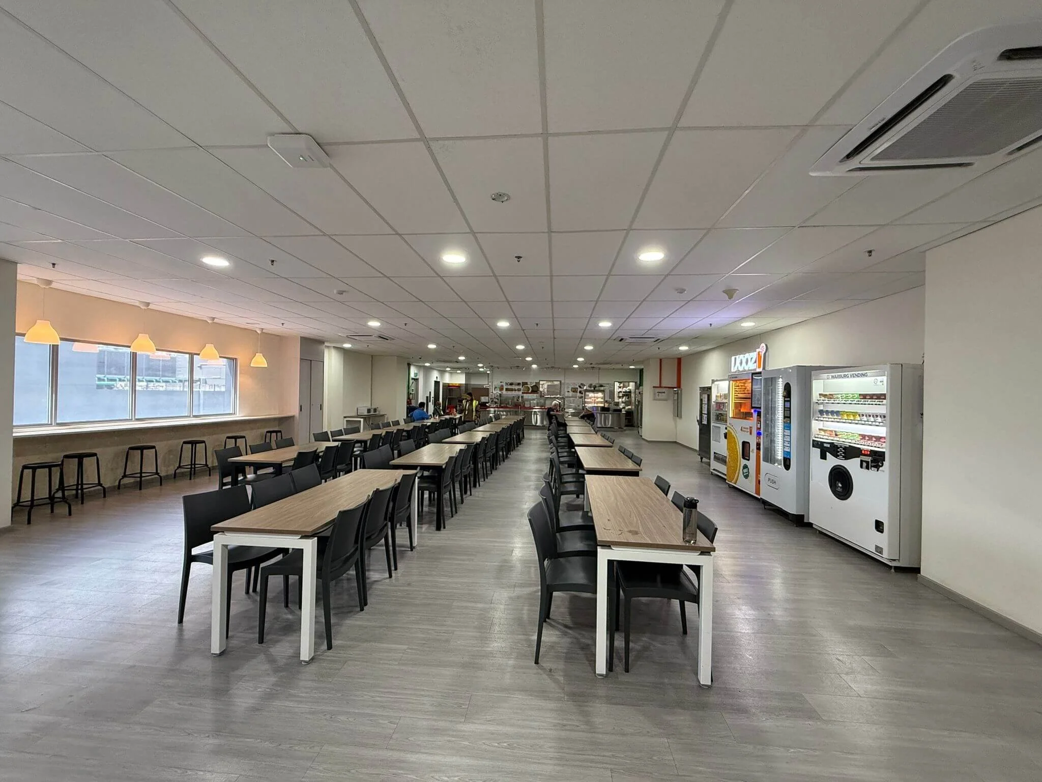 Empty cafeteria with long tables and chairs, vending machines on the right, and a window with hanging lights on the left.