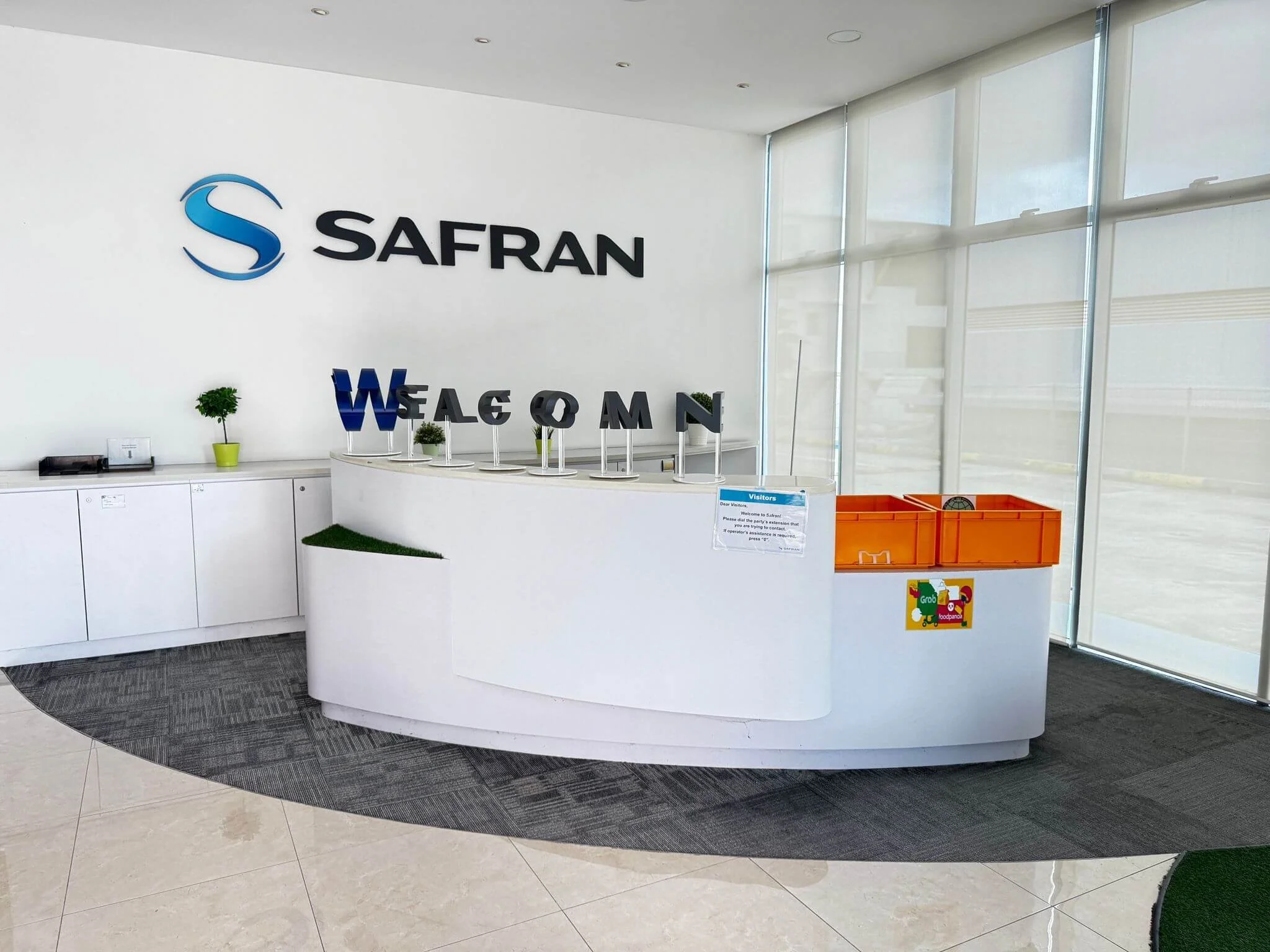 Reception desk with sign reading 'SAFRAN' above it, decorated with small potted plants and black letters spelling 'WELCOME'. There is a sign on the desk and orange boxes and a scale to the right, with large windows to the right providing natural ligh