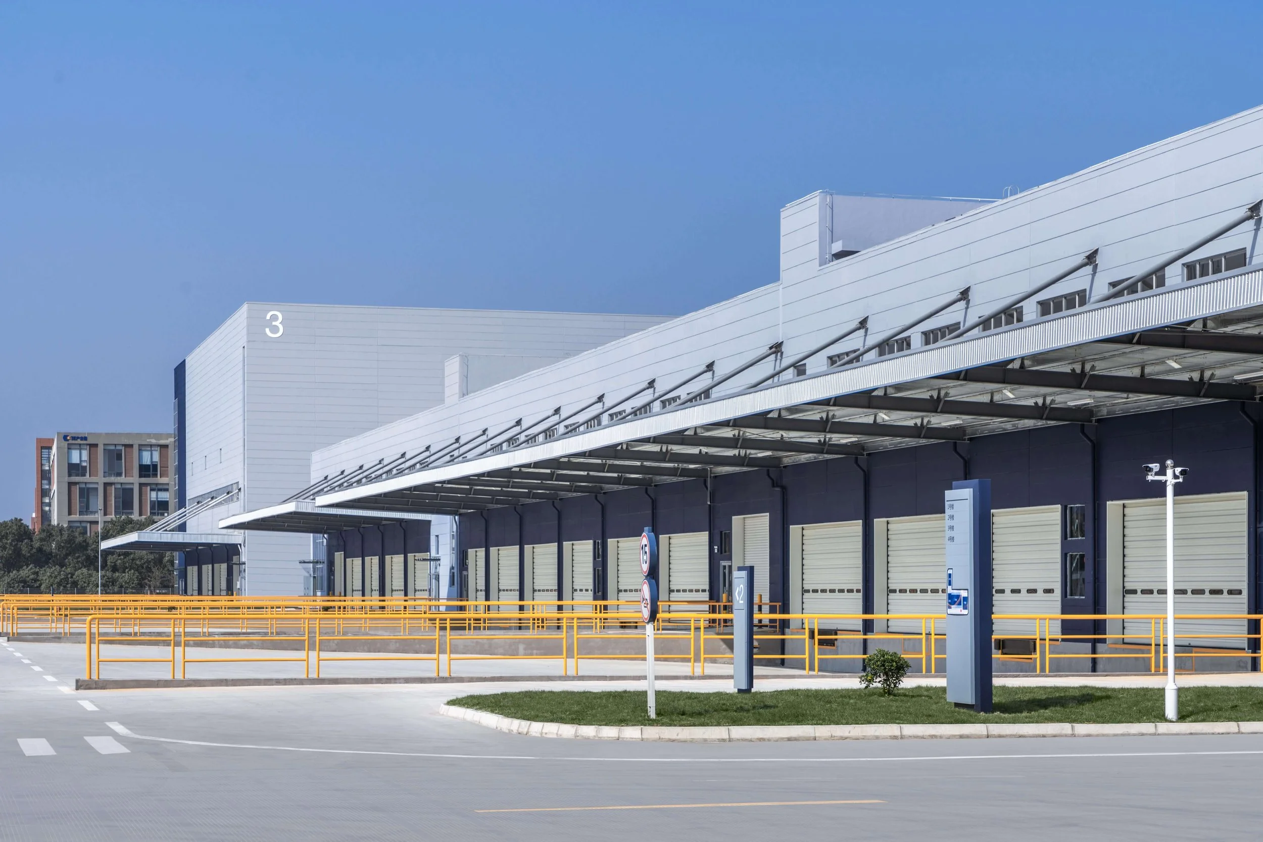 Large modern warehouse or distribution center with multiple loading docks, yellow safety barriers, and clear signage under a blue sky.