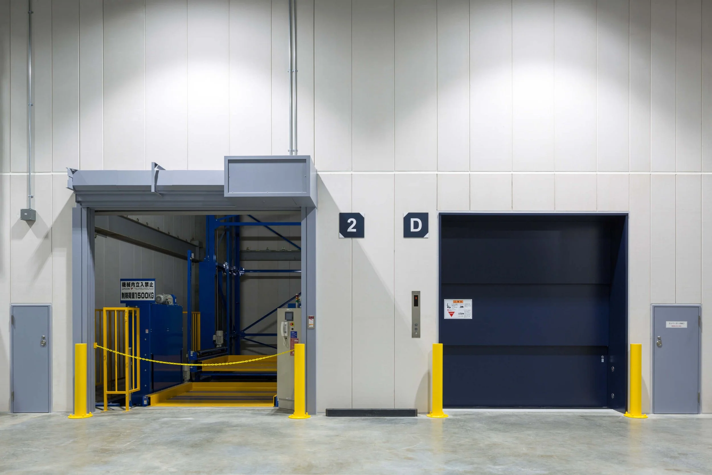 An industrial freight elevator and a car elevator located in a warehouse or loading dock area, with the freight elevator on the left and the car elevator on the right, both secured with yellow safety bollards.