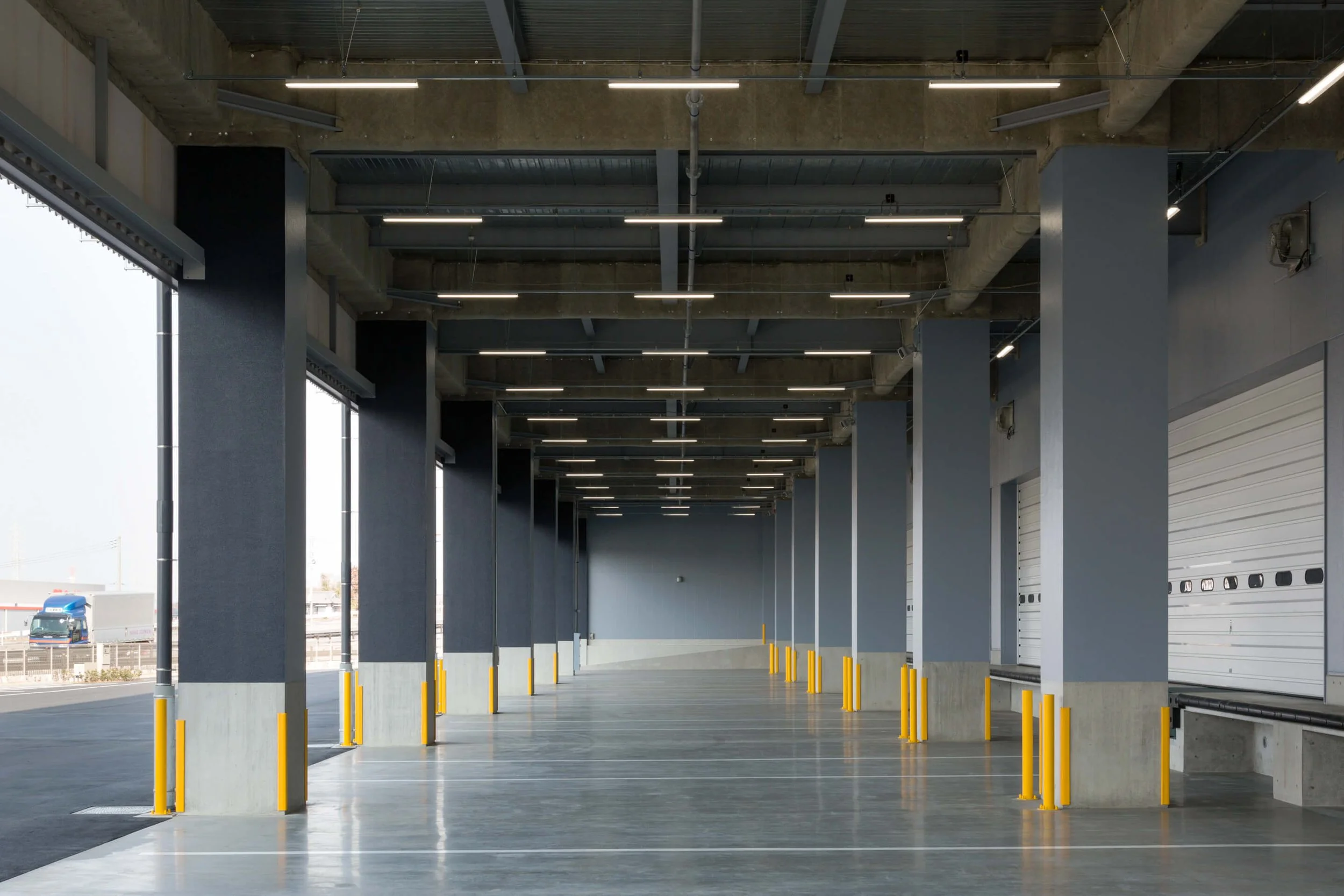 Empty loading dock with concrete floor, blue walls, yellow safety bollards, and overhead industrial lighting.