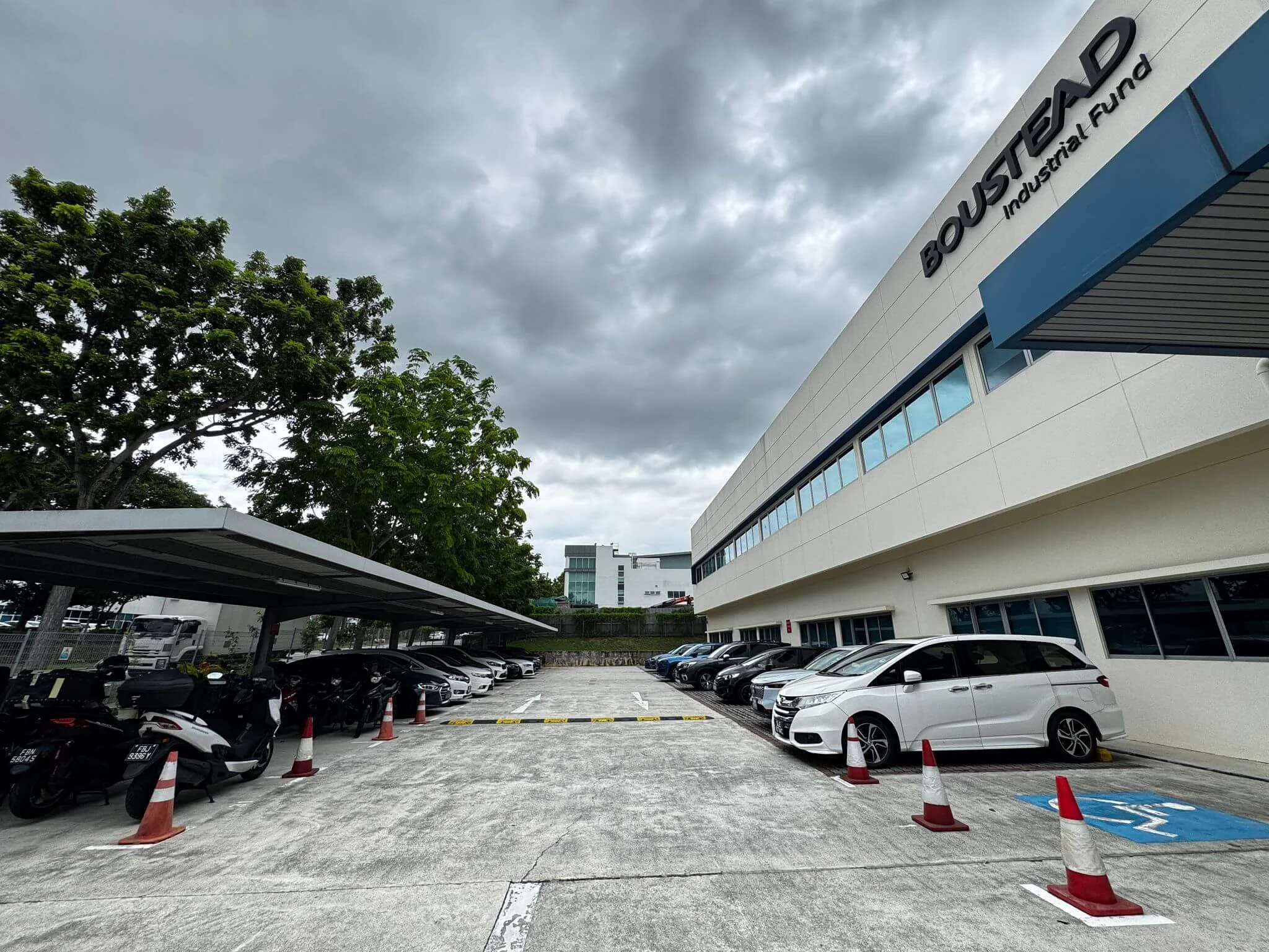Parking lot with cars and motorcycles in front of a white building with a sign that reads 'Bouwstad Industrial Fund' under a cloudy sky.