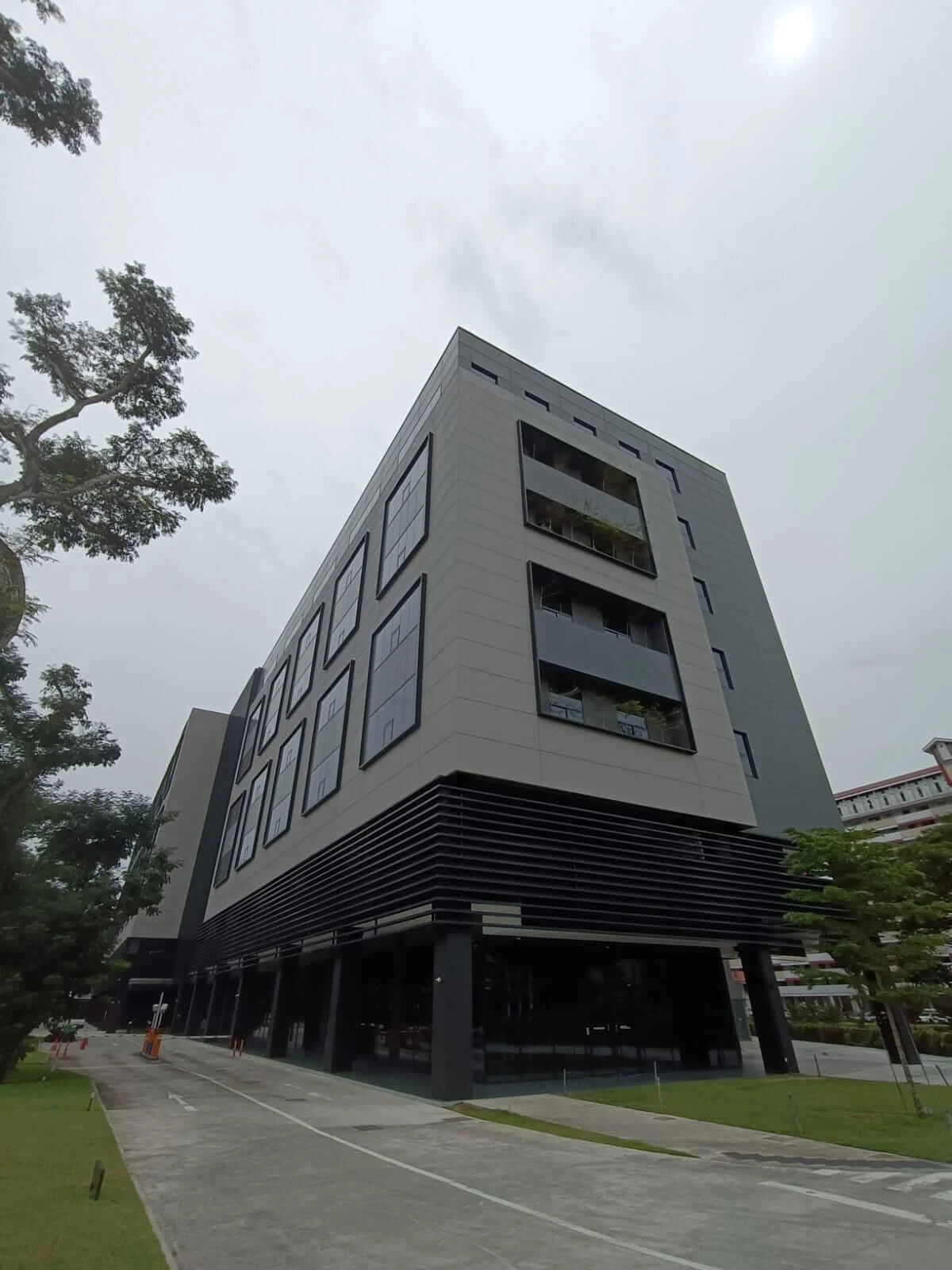Modern multi-story building with gray and black exterior, large windows, and a landscaped walkway in the foreground under cloudy sky.