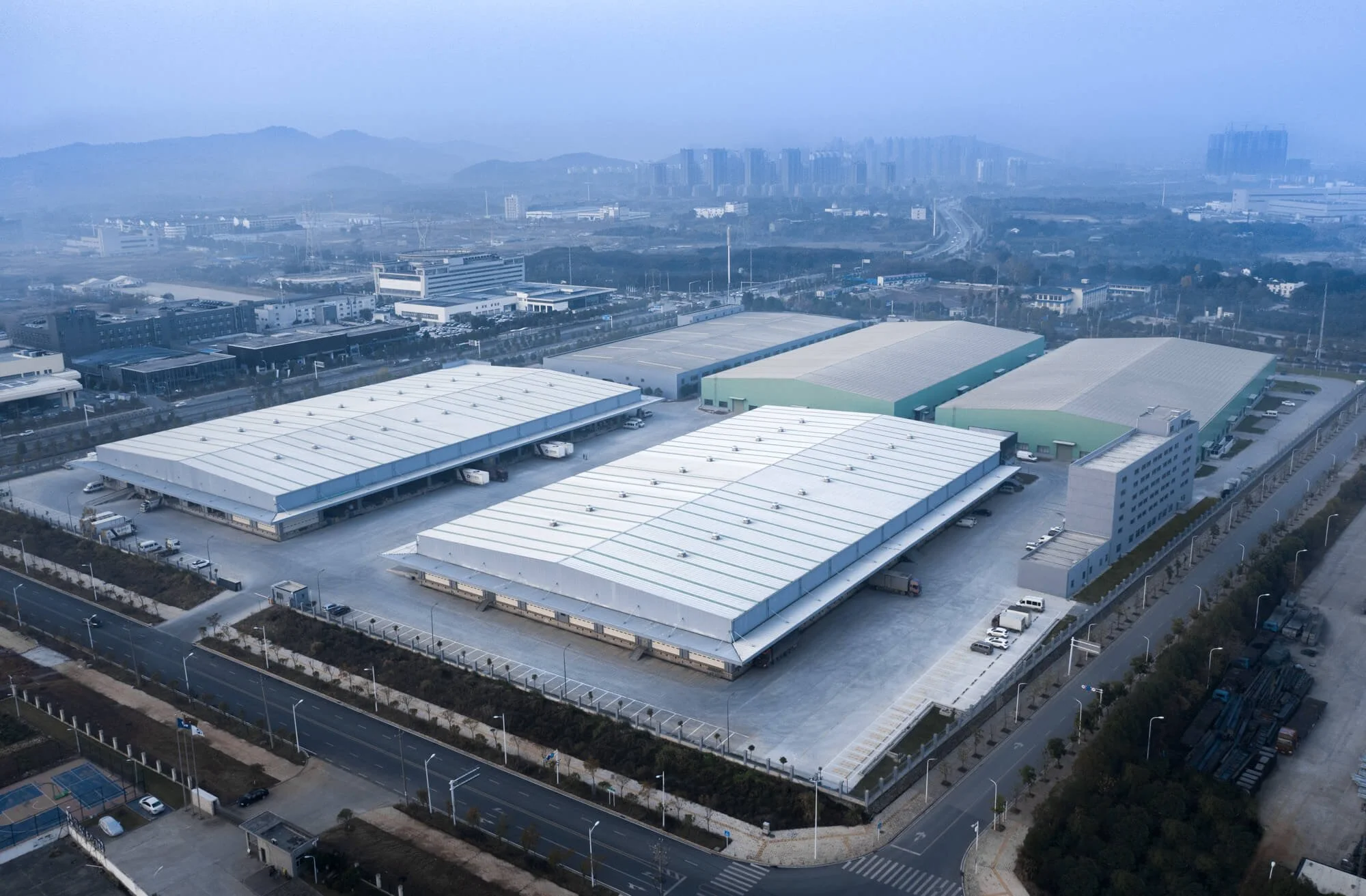 Aerial view of a large industrial warehouse complex with multiple white and green buildings, loading docks, parked trucks, and surrounding roads in an urban area with hills in the background.
