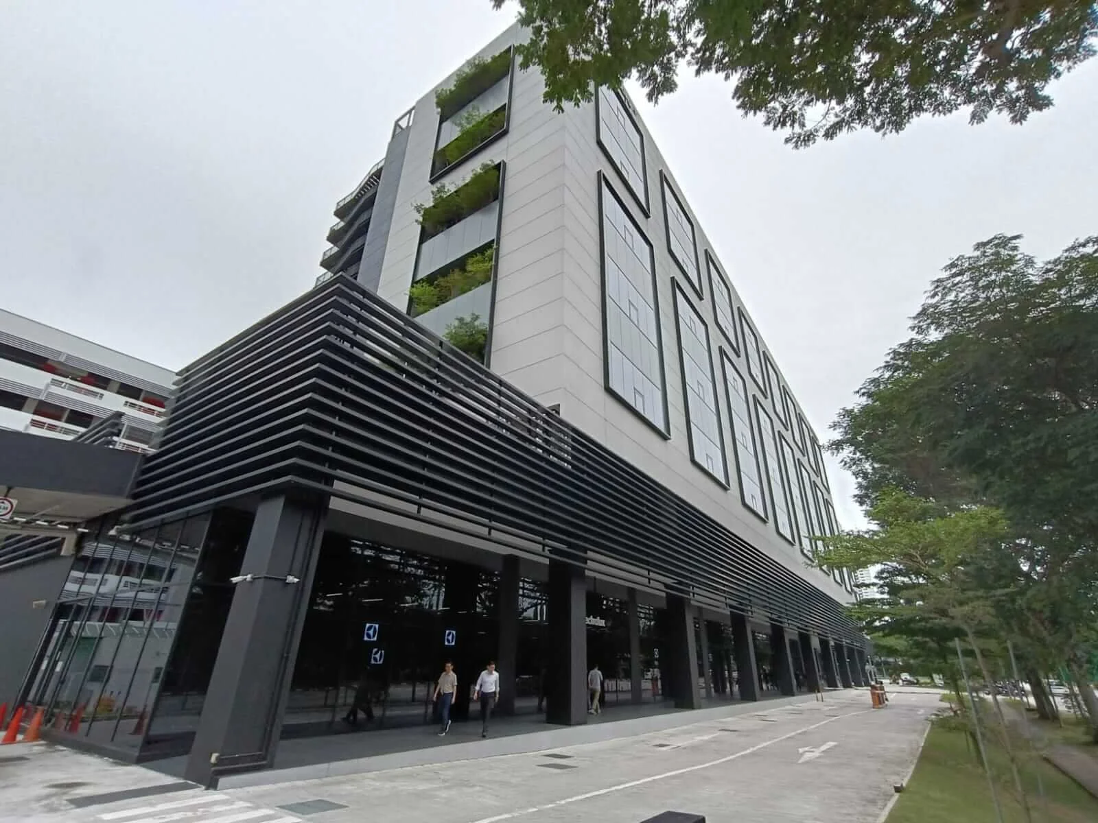 Modern multi-story building with black accents, large windows, and greenery on the upper balconies, located on a street with trees and pedestrians.