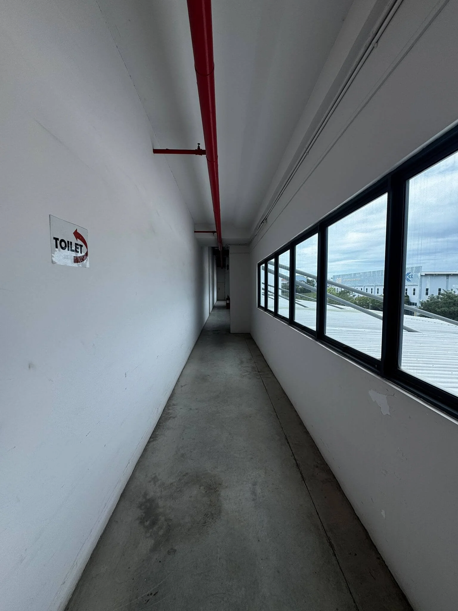 Empty indoor corridor with white walls, large windows on the right, red pipe running along the ceiling, and a sign pointing to the toilet on the left wall.