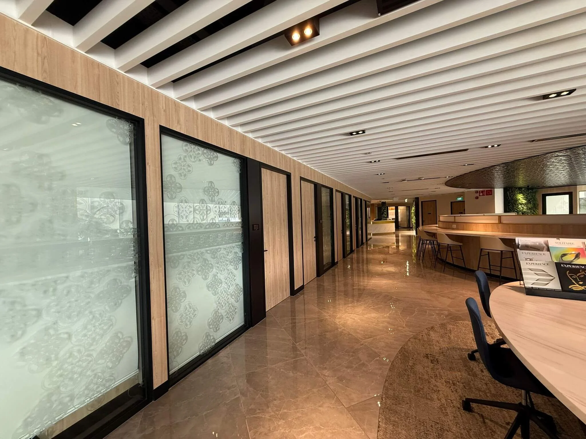 Modern interior space with wooden walls, glass panels, marble floor, and a curved bar area with black chairs, under a white slatted ceiling with recessed lighting.