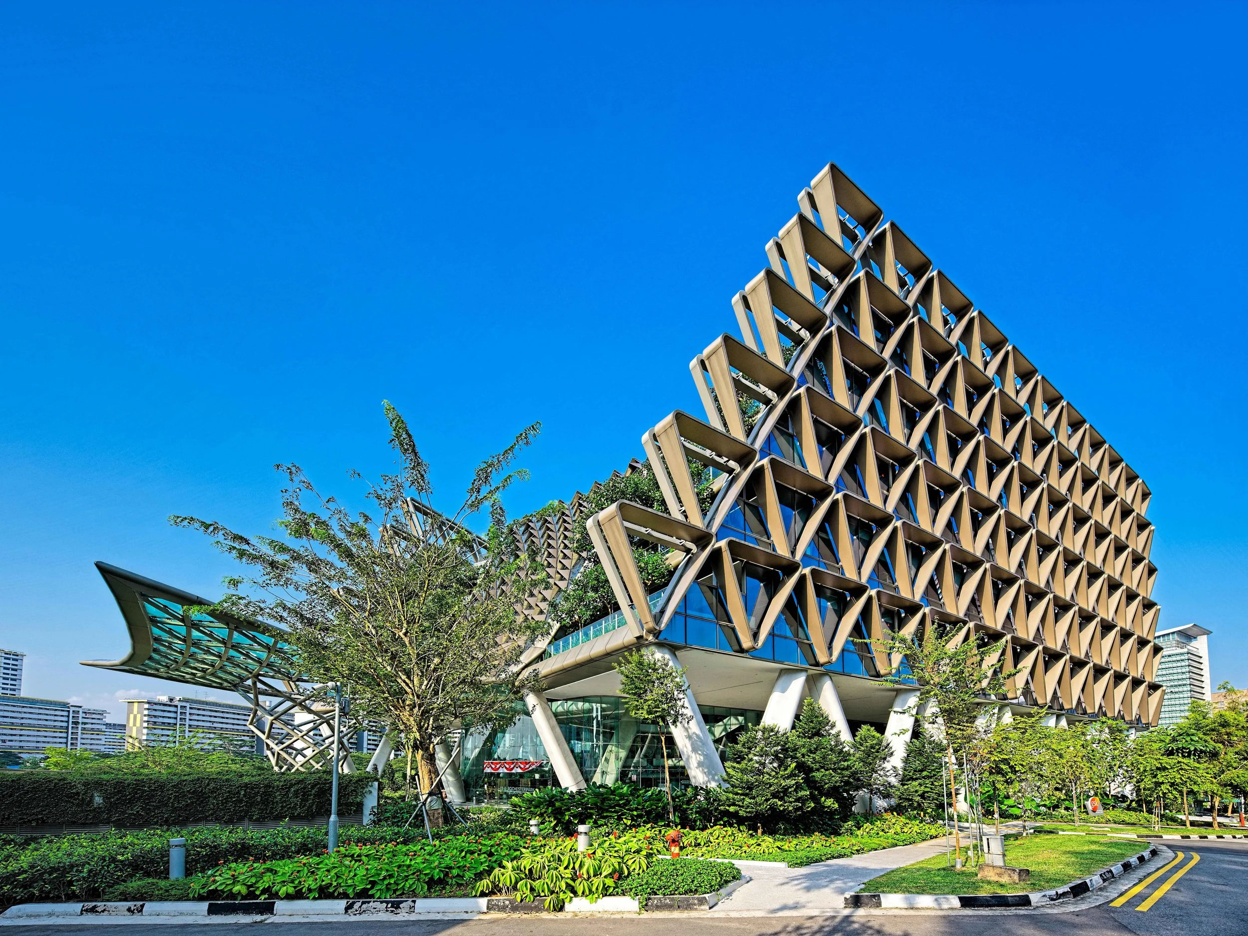 Modern building with a geometric facade made of metal and glass, surrounded by greenery, under a clear blue sky.