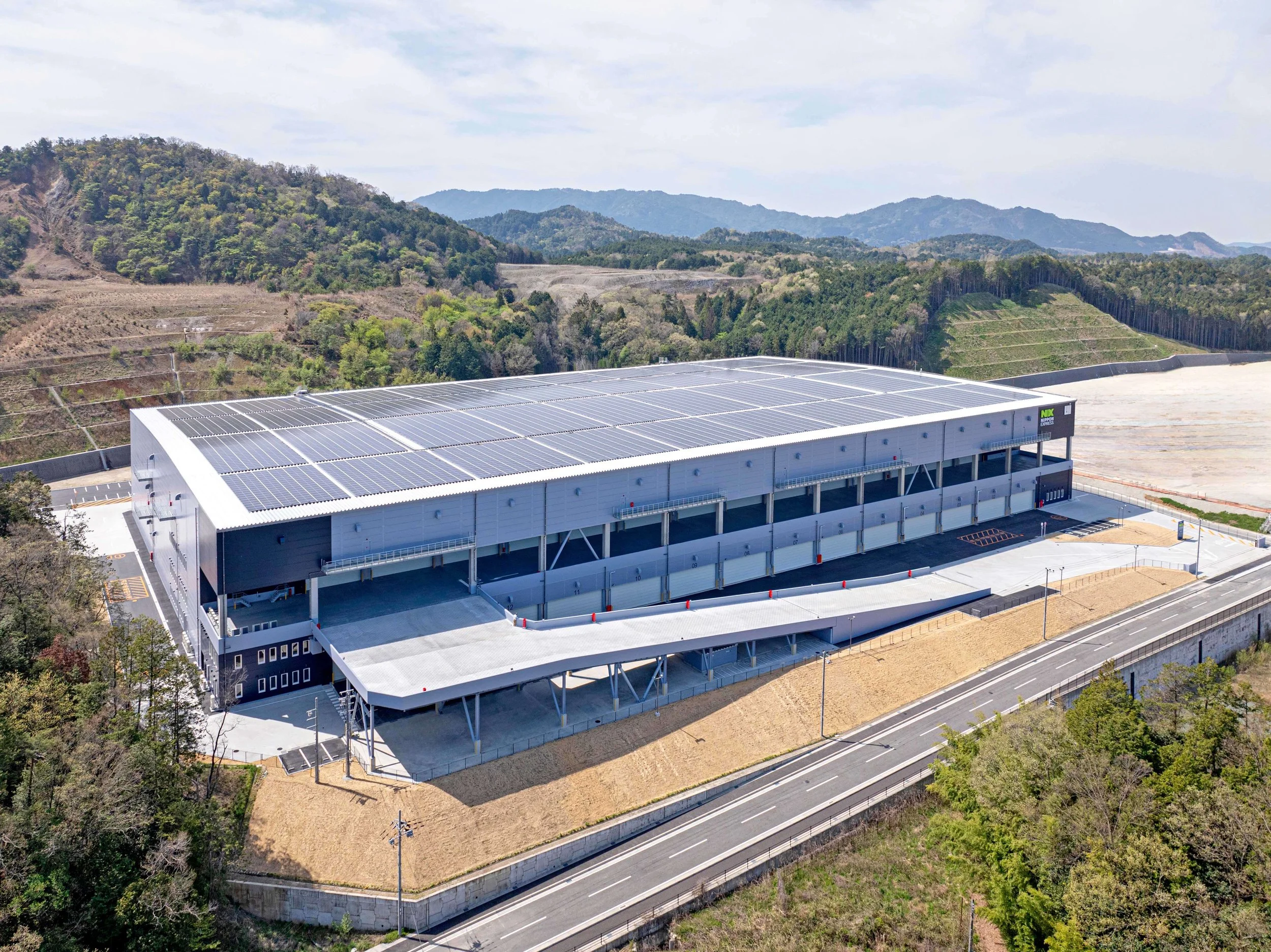 Large industrial building with a solar panel roof situated in a hilly, green landscape with mountains in the background.