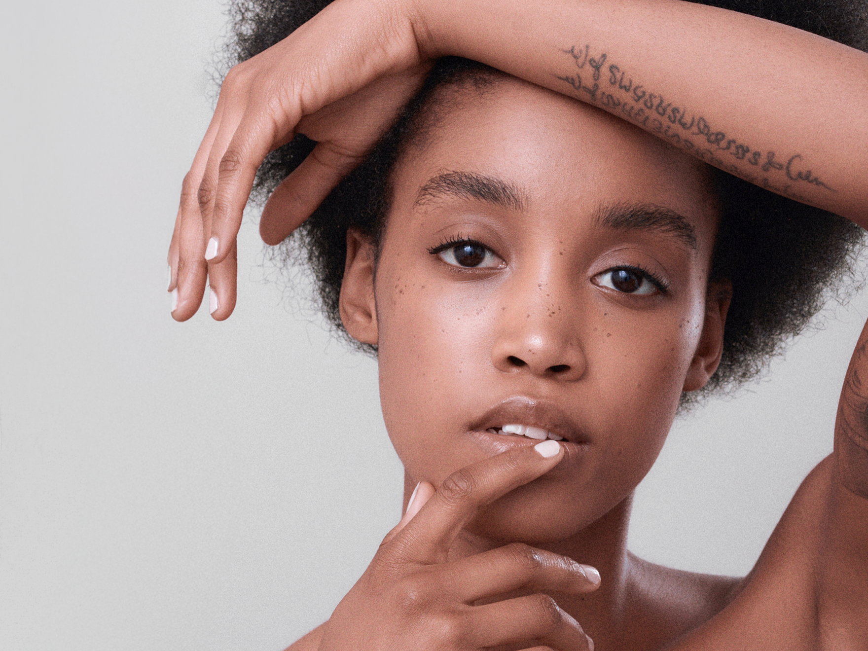 Two women with natural curly hair and freckles pose closely with contemplative expressions against a neutral background.