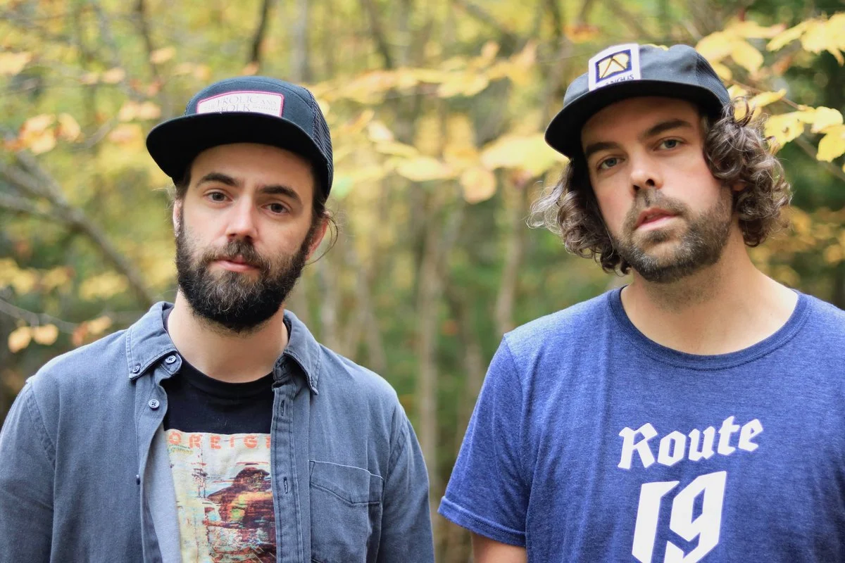 Two men standing outdoors with a forest background, both wearing baseball caps and casual t-shirts.