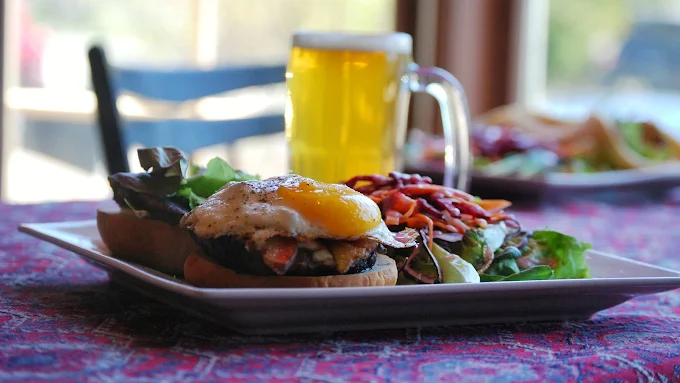 Plate with a burger topped with a fried egg, side salad, and a tall glass of beer on a red and purple patterned tablecloth.