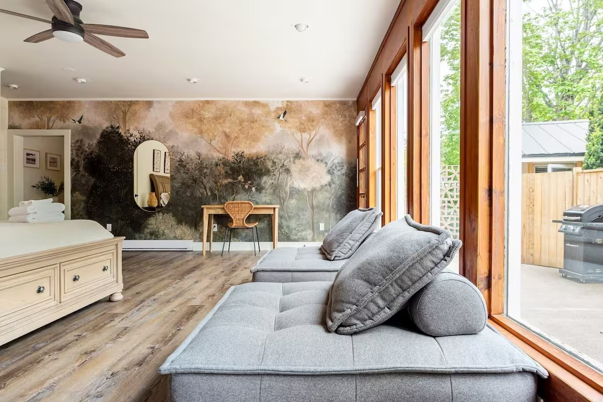 Living room with large windows, gray sofa with cushions, a desk with a wicker chair, a wall mural of trees, and a ceiling fan.