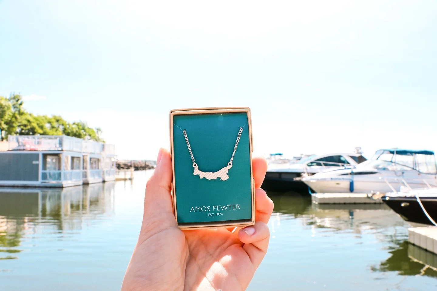 Hand holding a box with a necklace featuring a map of Ireland near a marina with boats and water in the background.