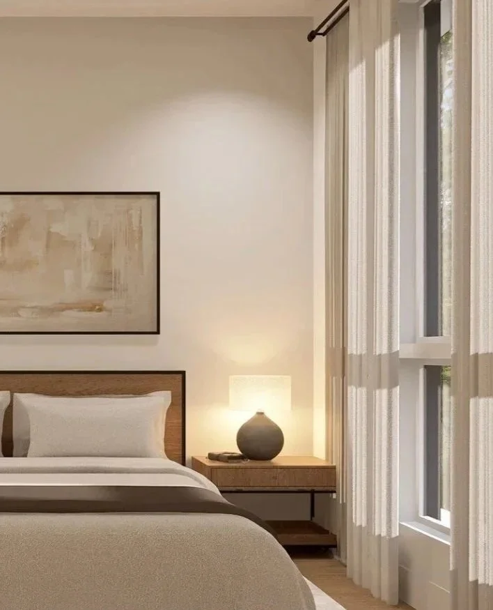 A cozy bedroom with a bed, a wooden nightstand with a lamp, and a window with white curtains.