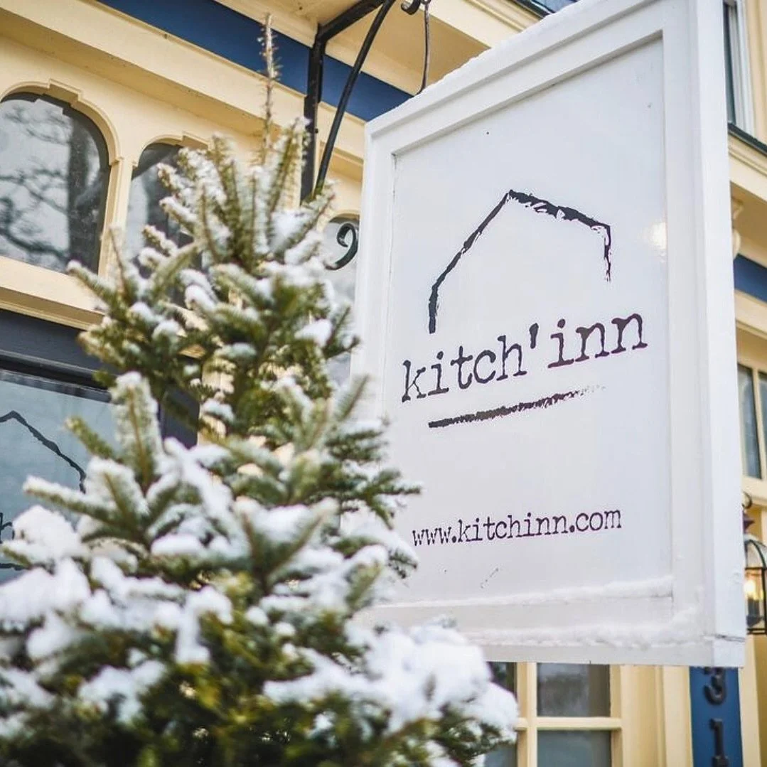 A white signboard with the text "kitch'inn" and a website link "www.kitchinn.com" is hanging outside a building. A small snow-covered evergreen tree is in the foreground.