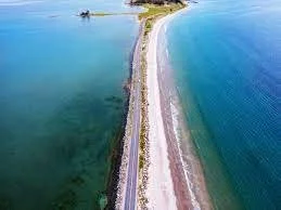 A narrow strip of land separates a body of water from the ocean, with a road running along the top edge and some greenery in the distance.