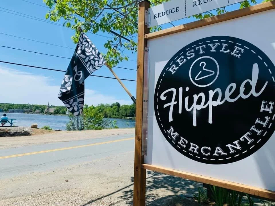 A sign for Flipped Marketplace, a resale store, is displayed outdoors next to a river. The sign has a circular logo with the words "ReSTYLE" and "Marketplace," and the store name "Flipped" in the center. A black and white patterned flag is waving nearby, and there are people sitting by the river in the background. The scene is bright and sunny, with green trees and clear blue sky.