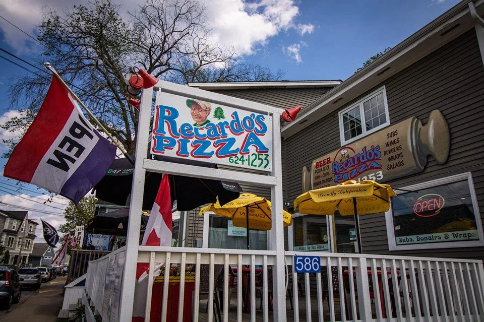 Exterior view of Recardo's Pizza restaurant with signage, yellow umbrellas, and flags, on a street with residential buildings in the background.