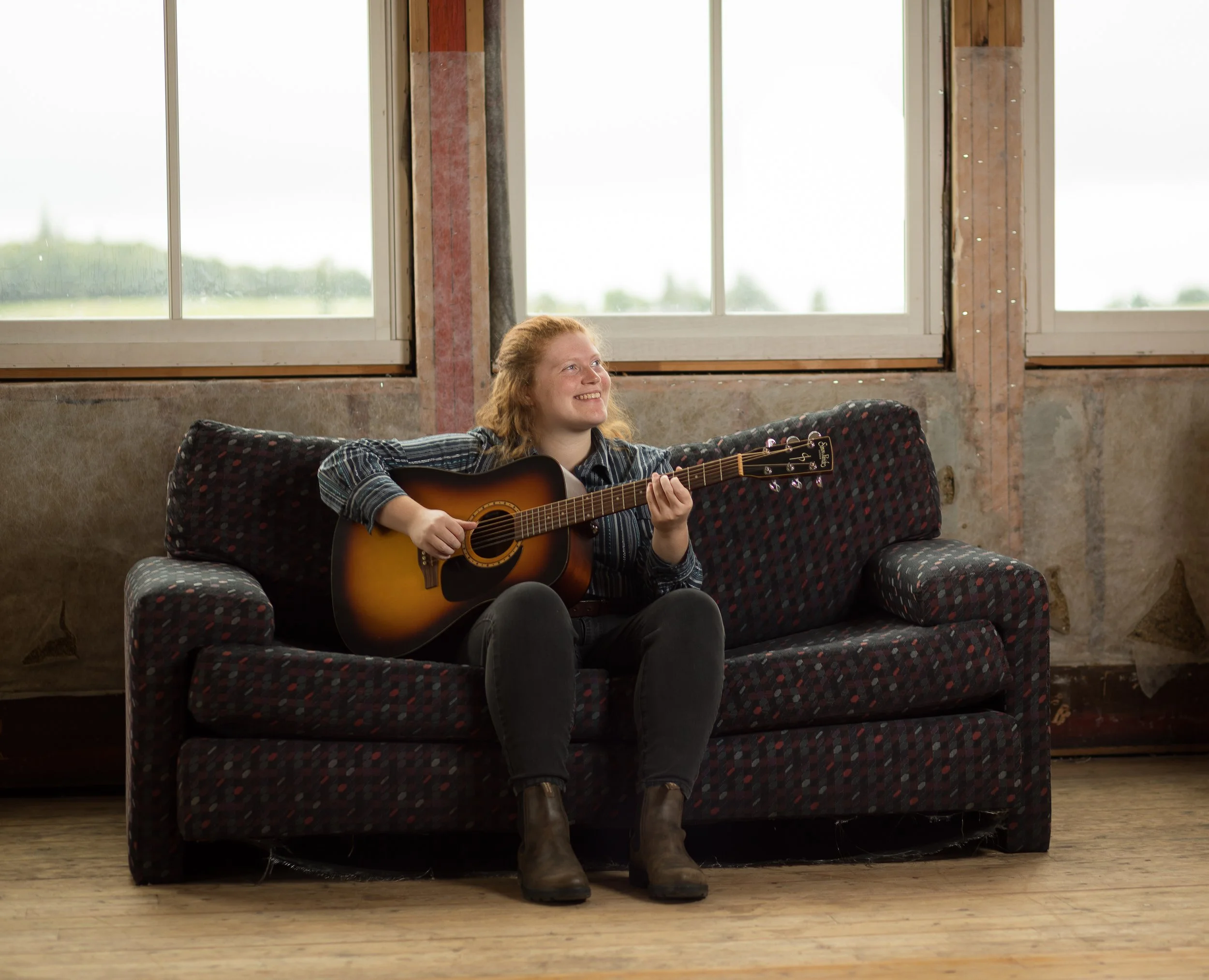 A young woman with red hair sitting on a patterned sofa, playing an acoustic guitar and smiling, in a rustic room with large windows and wooden floors.