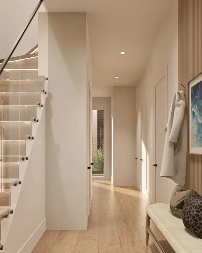 Bright hallway with white walls, wooden flooring, a staircase with glass railing on the left, a bench with cushions on the right, and closed white doors. There is also a coat hanging on a hook and a painting on the wall.