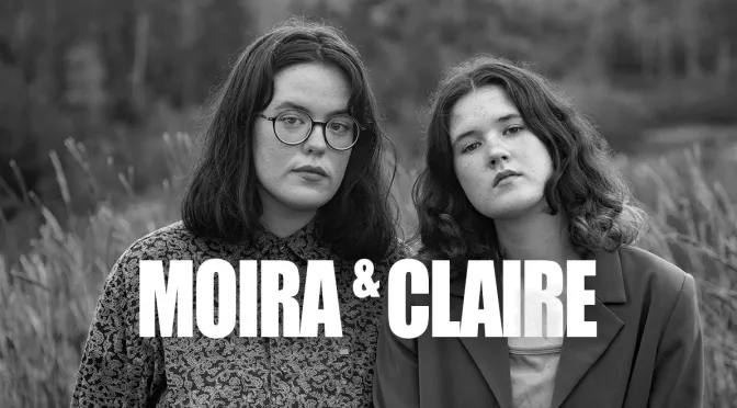 Black and white photo of two young women, Moira and Claire, standing outdoors with blurred foliage in the background. Moira has shoulder-length dark hair, glasses, and wears a patterned top. Claire has curly hair and wears a dark blazer. Text overlay reads "MOIRA & CLAIRE" in bold white letters.