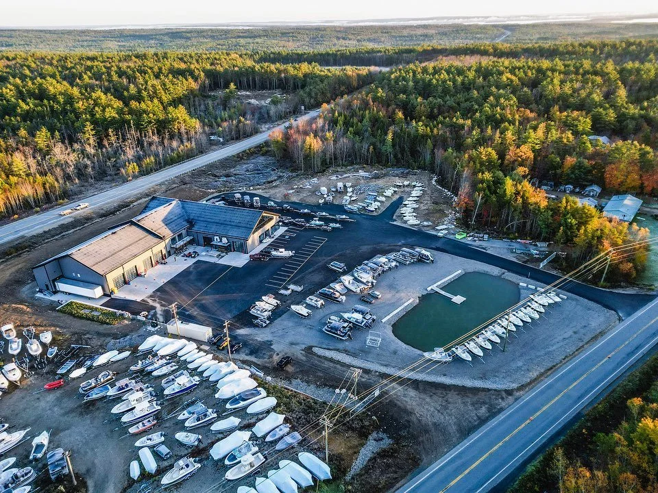 Aerial view of a marina with boats, a large building, parking lot, and a swimming pool, surrounded by trees and nearby roads.