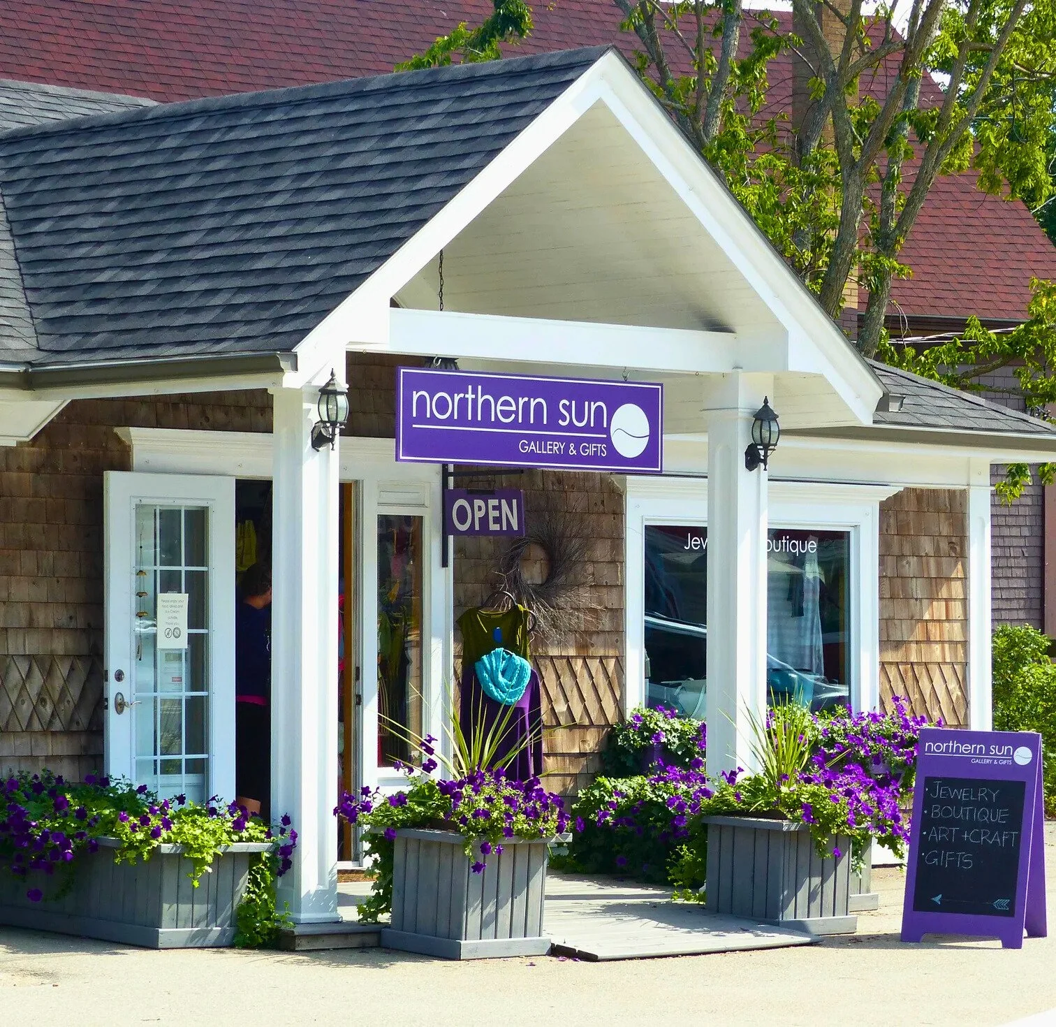 A small boutique storefront called 'Northern Sun Gallery & Gifts' with purple signage, flower planters with purple flowers, signs indicating it is open, and a chalkboard sign listing jewelry, boutique, art and craft, and gifts.