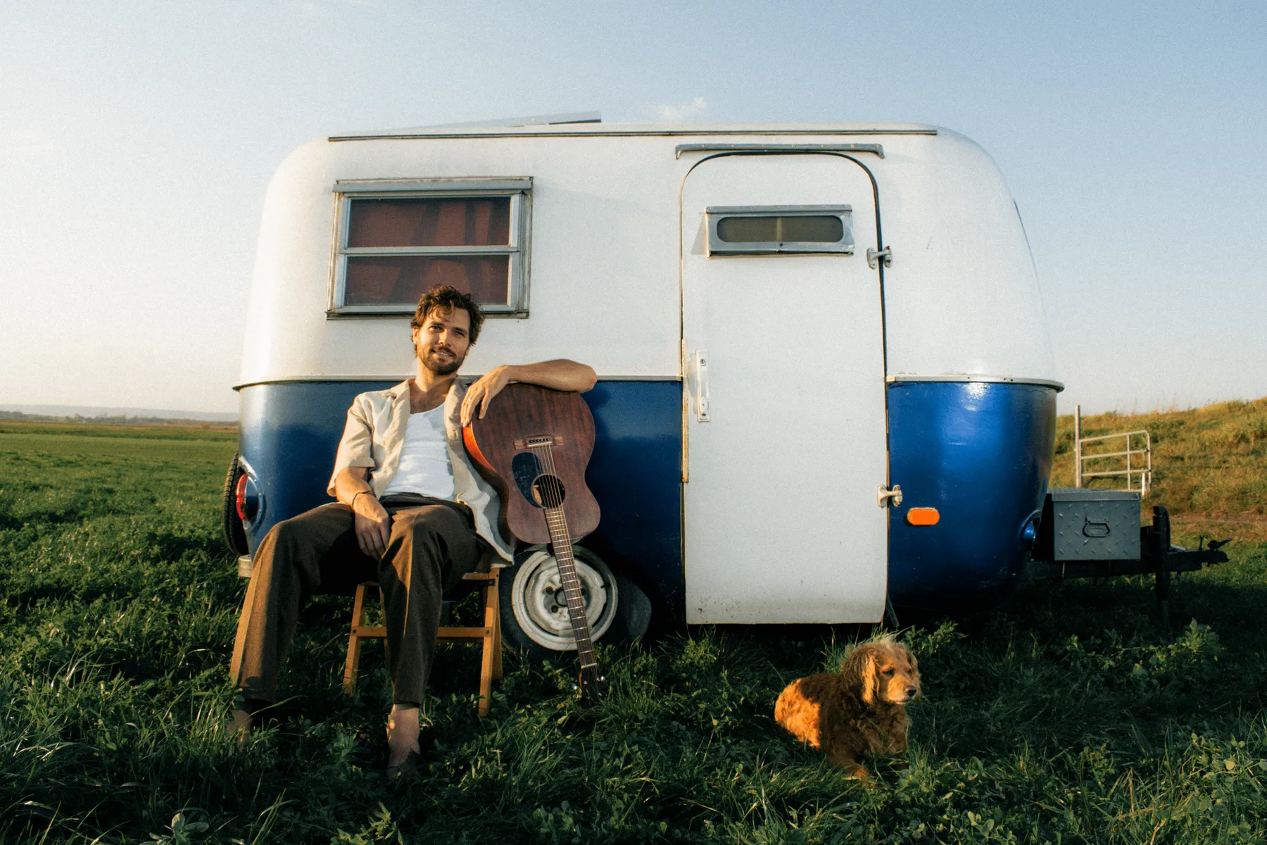 A man sitting on a wooden chair next to a vintage camper trailer, with a guitar resting against the trailer and a small dog sitting on the grass nearby, in a grassy field during late afternoon or early evening.