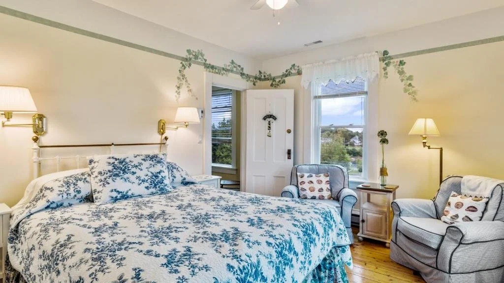 Cozy bedroom with a white bed featuring blue floral bedding, two armchairs with patterned pillows, and a window showing a view of trees and sky.
