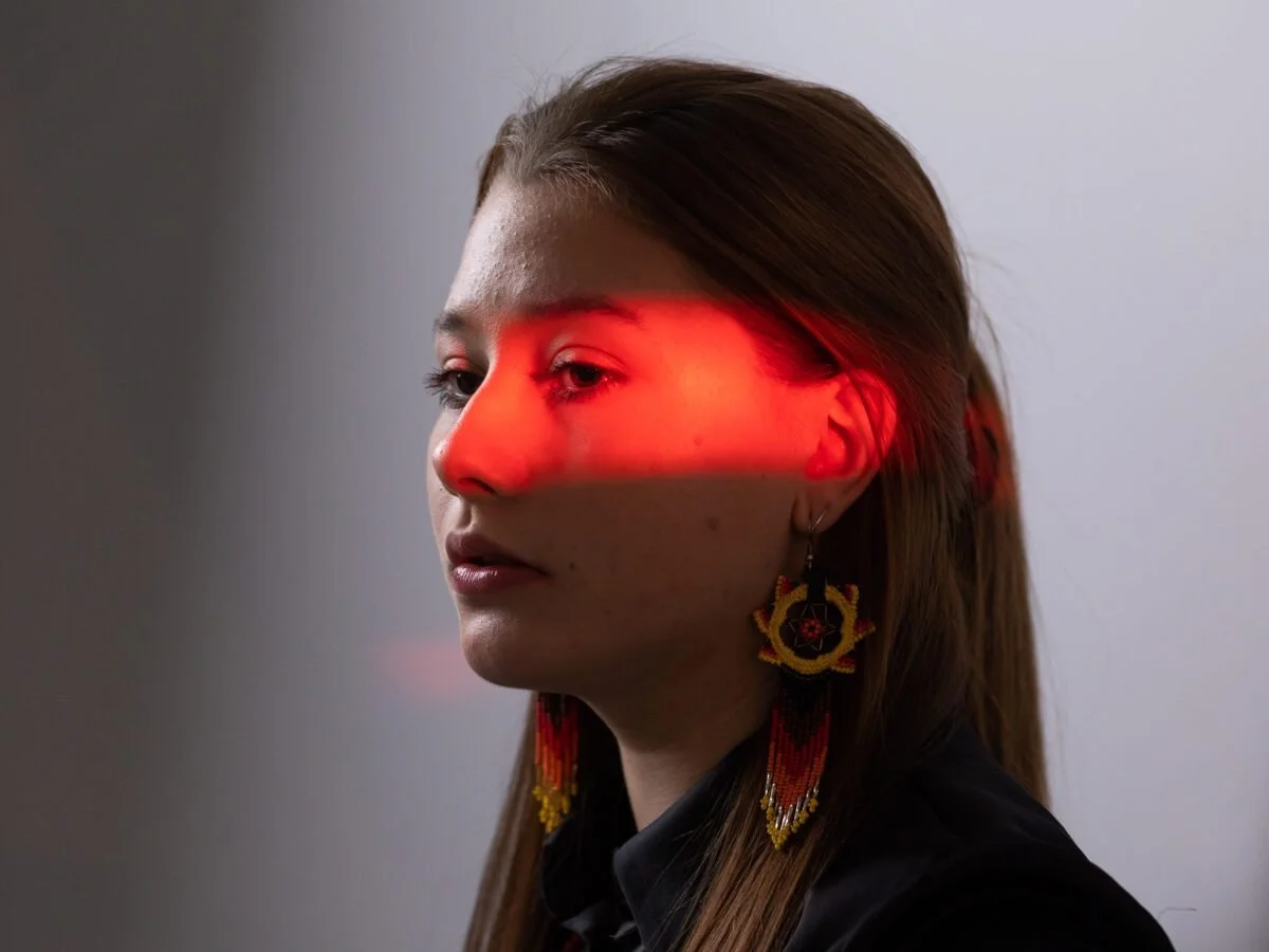 Side profile of a woman with long brown hair and colorful beaded earrings, with a red laser beam across her face and eye.