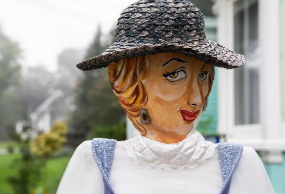 A garden gnome dressed as a woman with painted facial features, wearing a black hat and a white dress with blue accents, standing outdoors in a residential area.