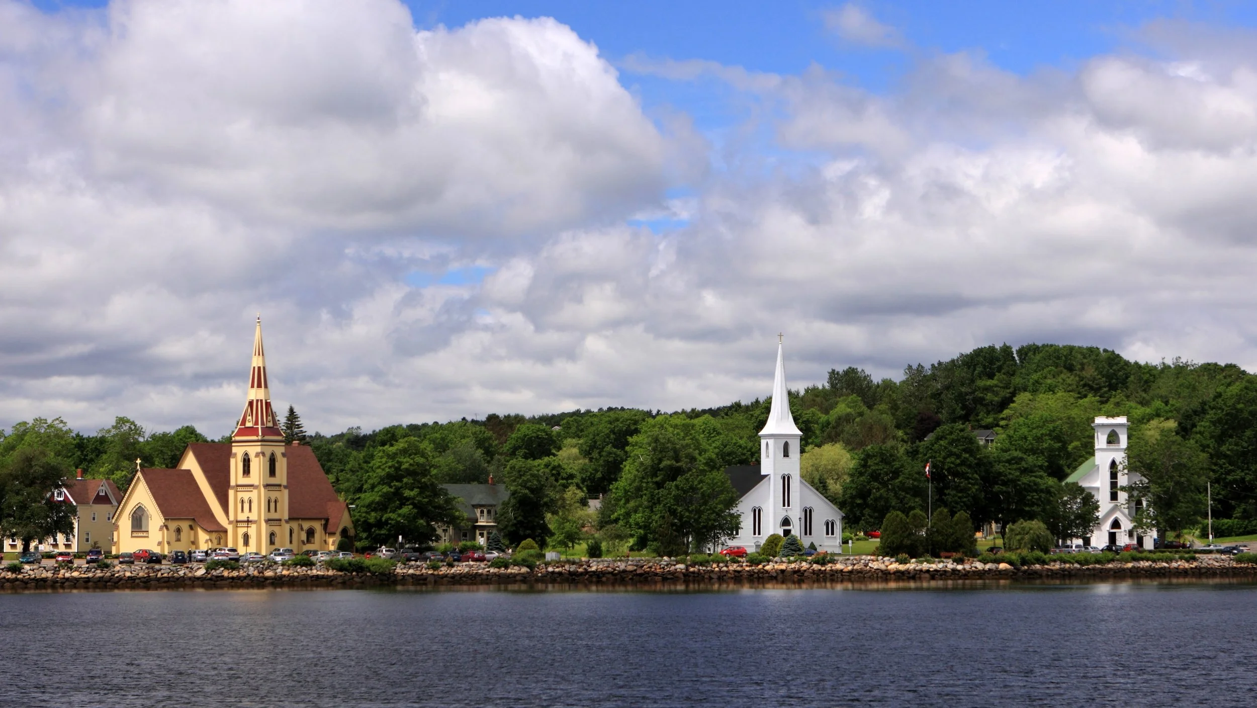 A lakeside view with two churches, one yellow with a tall steeple and the other white with a pointed steeple, surrounded by green trees and a partly cloudy sky.