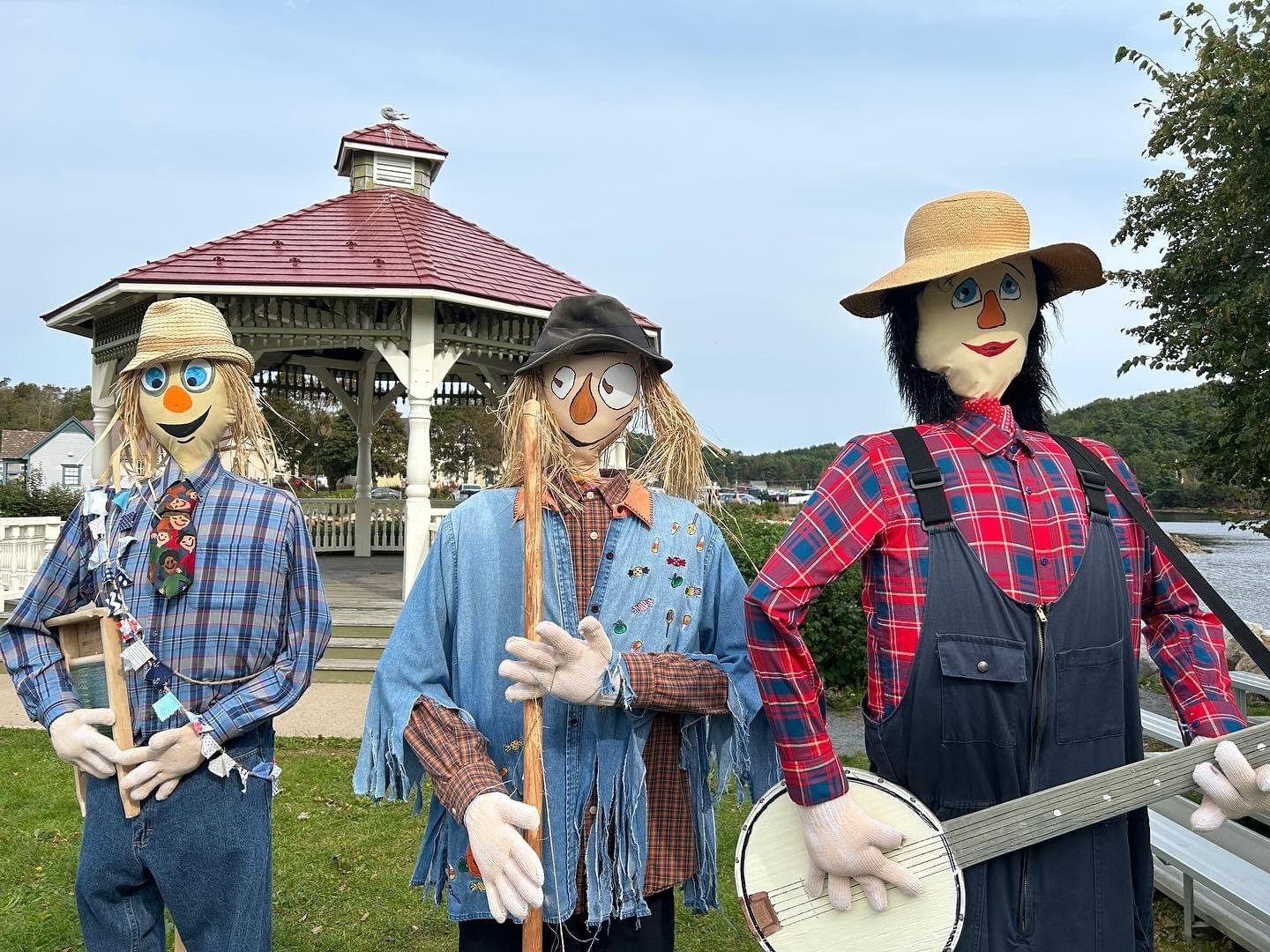 Three scarecrow figures dressed in plaid, denim, and overalls with straw and fabric faces, wearing hats, standing outdoors near a gazebo and water. One holds a small wooden sign, another holds a stick, and the third holds a banjo.