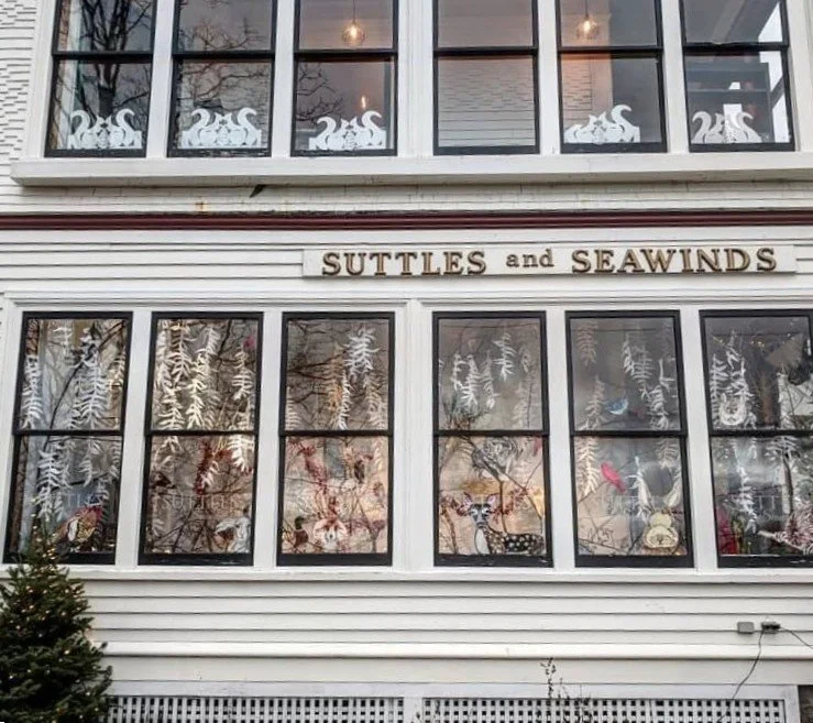 Storefront with the sign "SUTTLES and SEAWINDS" above large windows decorated with white paper cutouts of leaves and animals, including birds and deer.