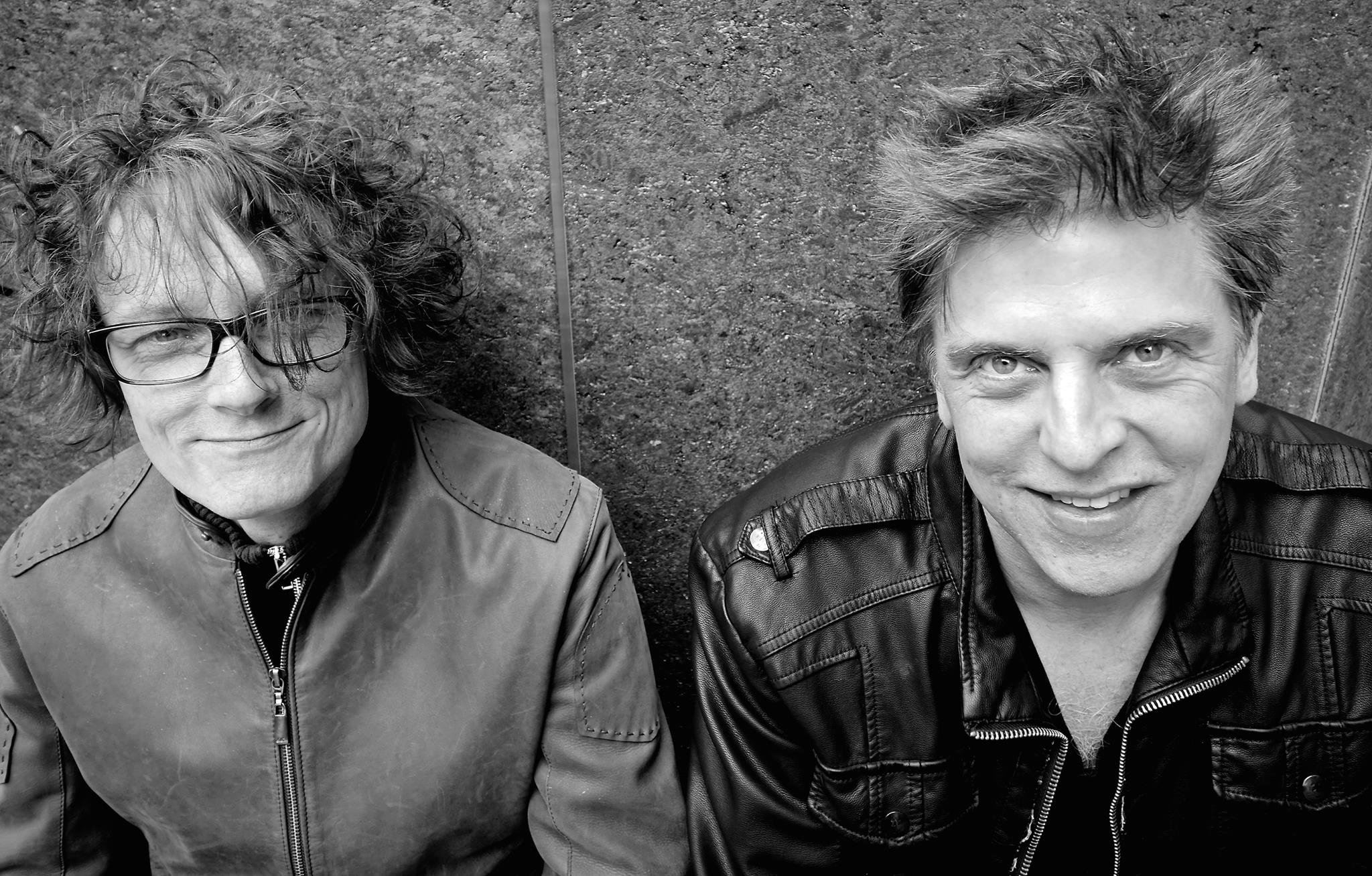 Black and white photo of two men sitting against a textured wall. The man on the left has curly hair, glasses, and is wearing a jacket. The man on the right has spiky hair and is dressed in a leather jacket.
