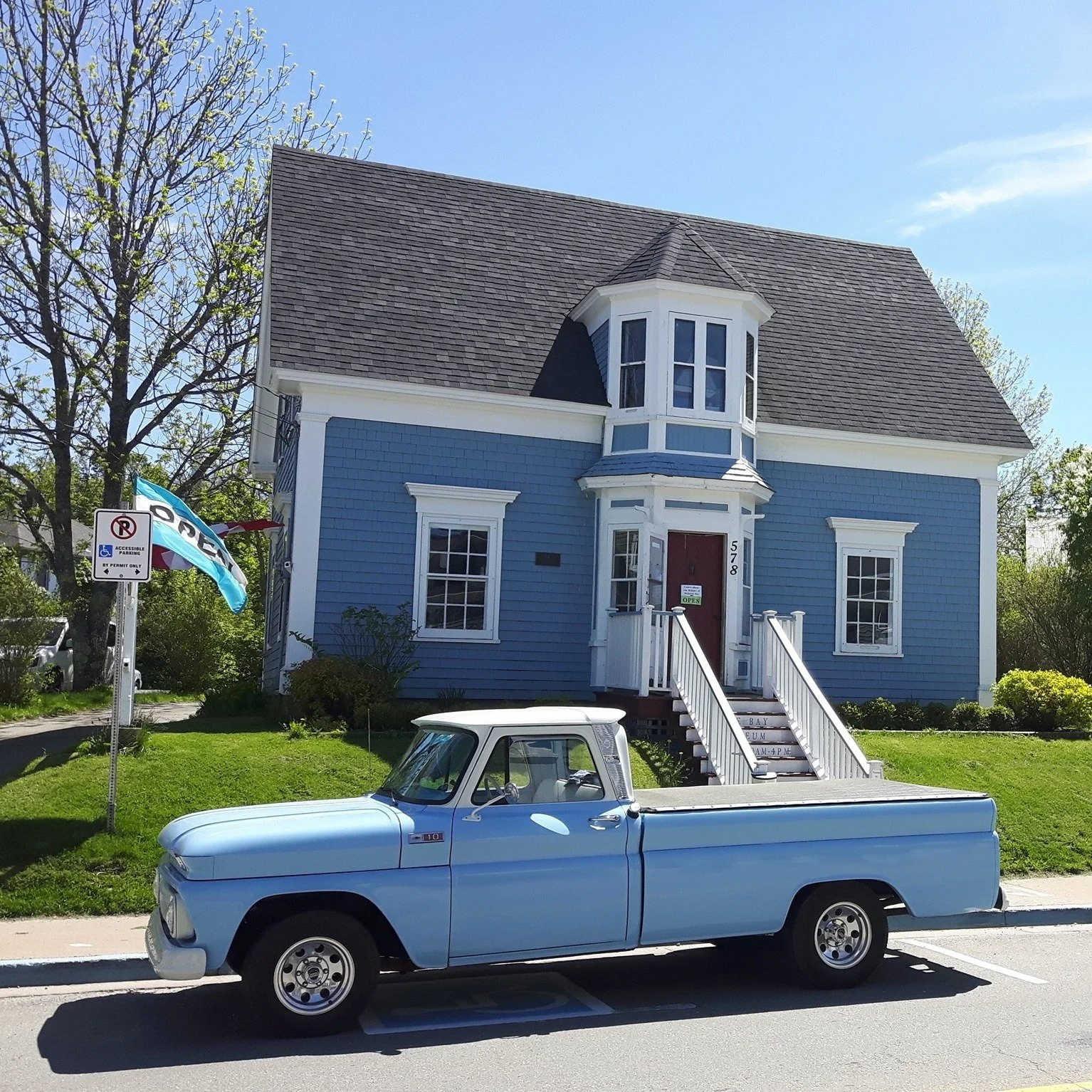 A blue house with white trim and a bay window, a small front porch with stairs, and a vintage blue pickup truck parked in front. A flag and a no parking sign are visible near the house.