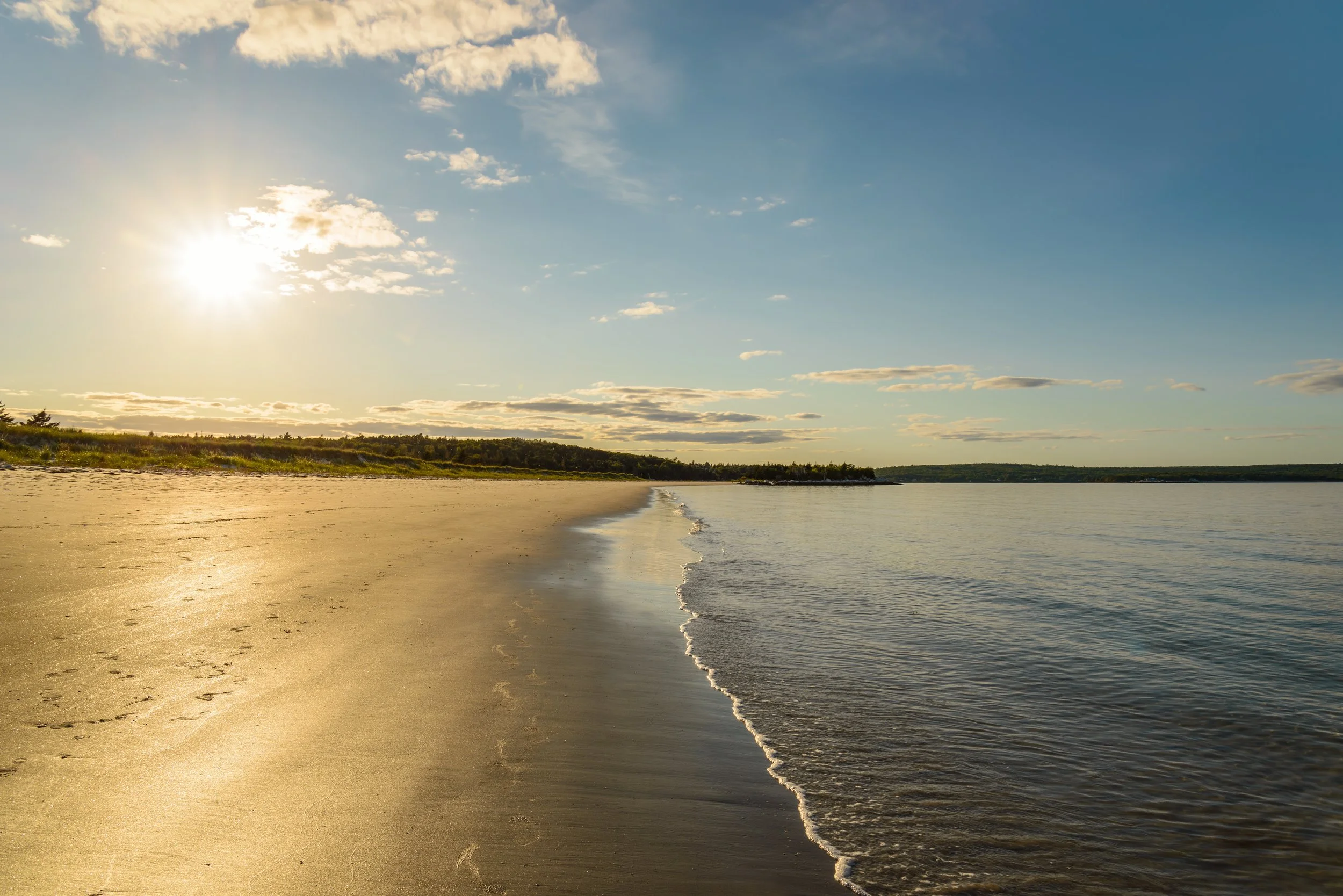 A calm beach with golden sand leading to the water, and a bright sun in the sky with some clouds, during sunset or sunrise.