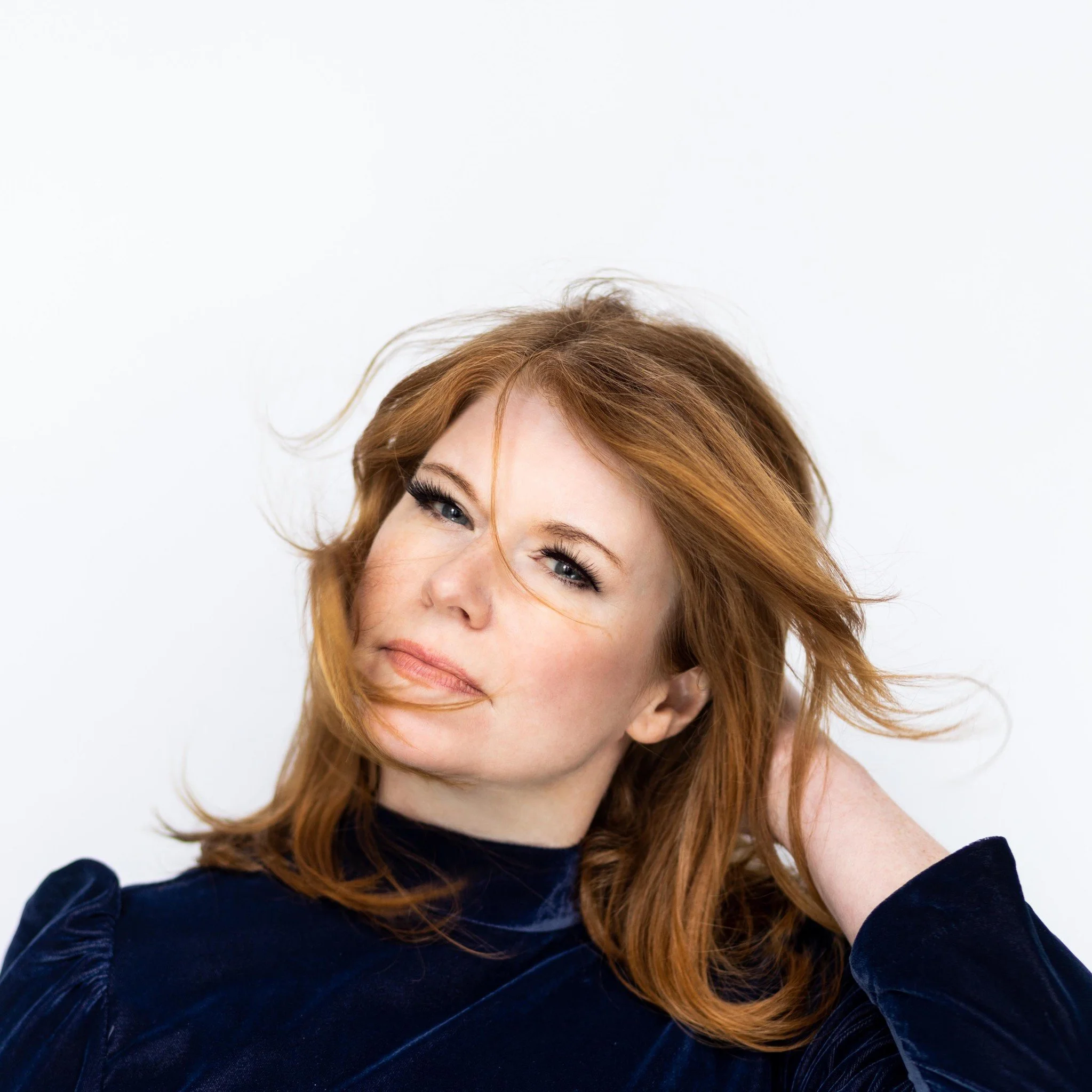 Close-up portrait of a woman with red hair, blue eyes, and fair skin, wearing a dark blue velvet top, posing with one hand behind her head against a plain white background.