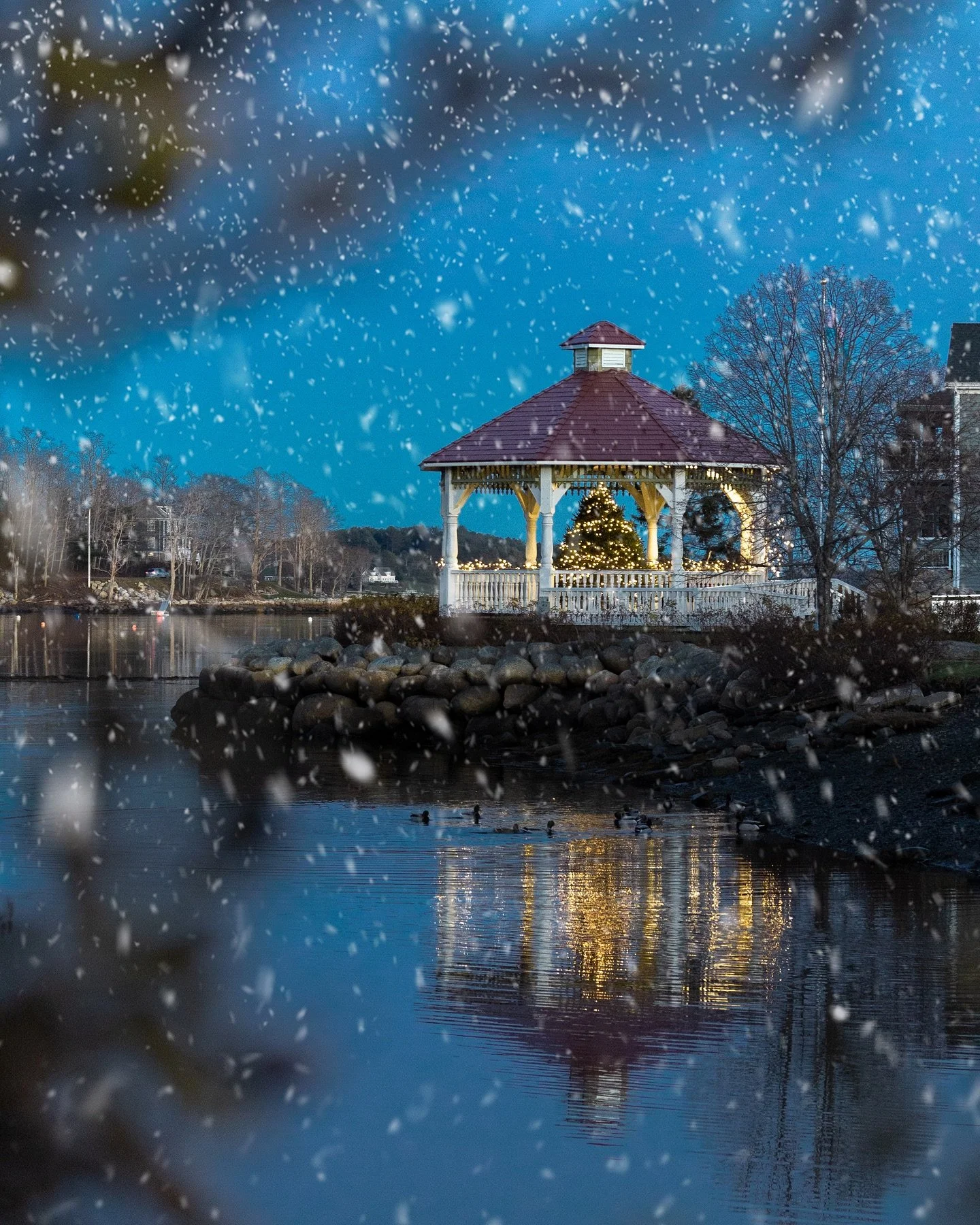 A gazebo with a decorated Christmas tree inside, seen through falling snow near a body of water at dusk.