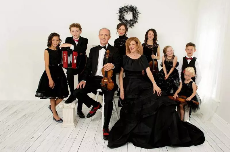 A family dressed in formal black attire, posing with violins and an accordion in a bright room with white walls and wooden floor.