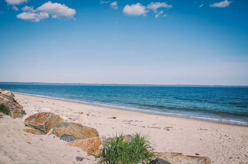 Sandy beach with large rocks and green plants in the foreground, blue ocean water, and a partly cloudy sky with a few small white clouds.