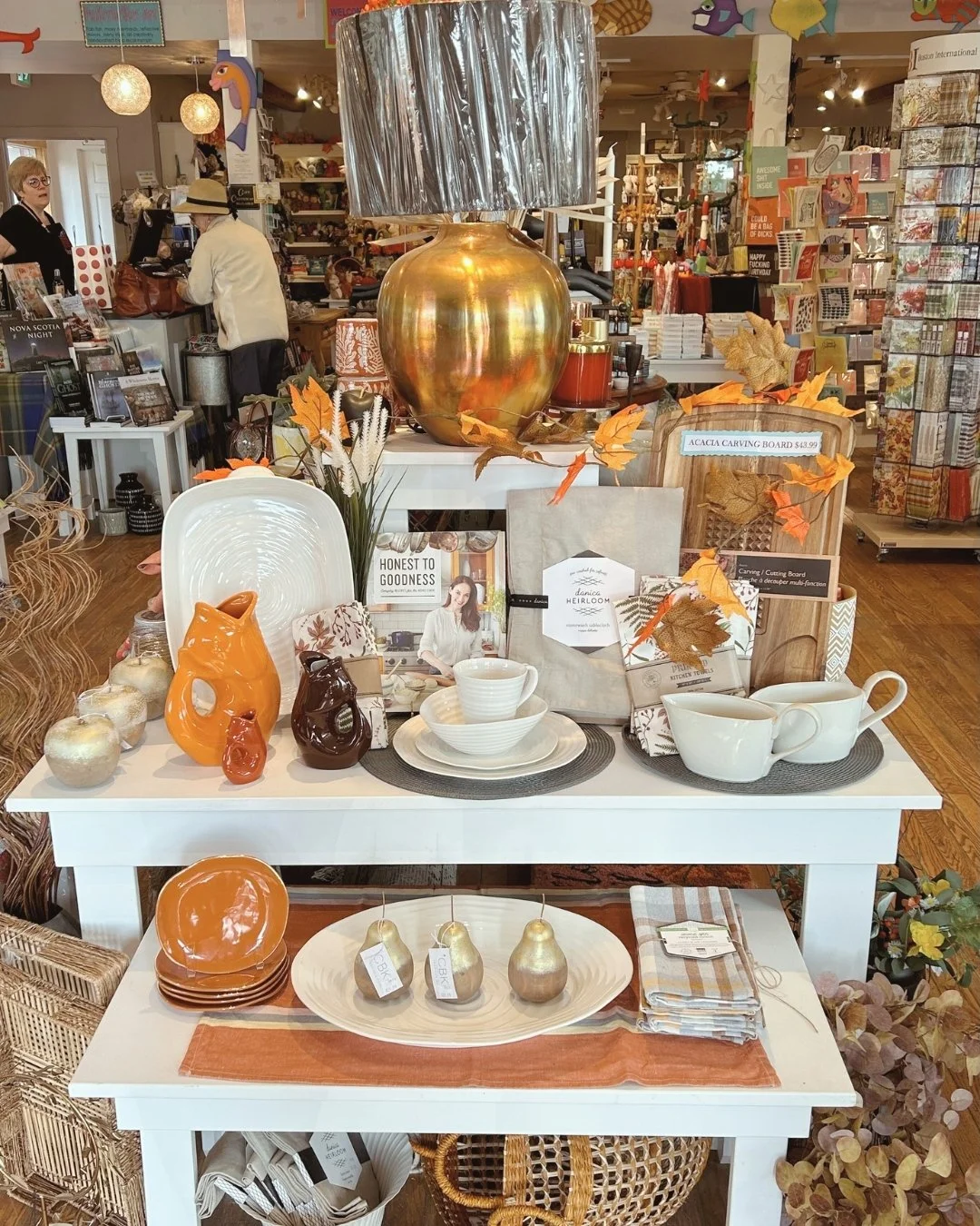 Display table filled with autumn-themed home decor, including ceramics, candles, plates, bowls, and books, with fall leaves and a large gold vase with a silver lampshade on top in a boutique store.