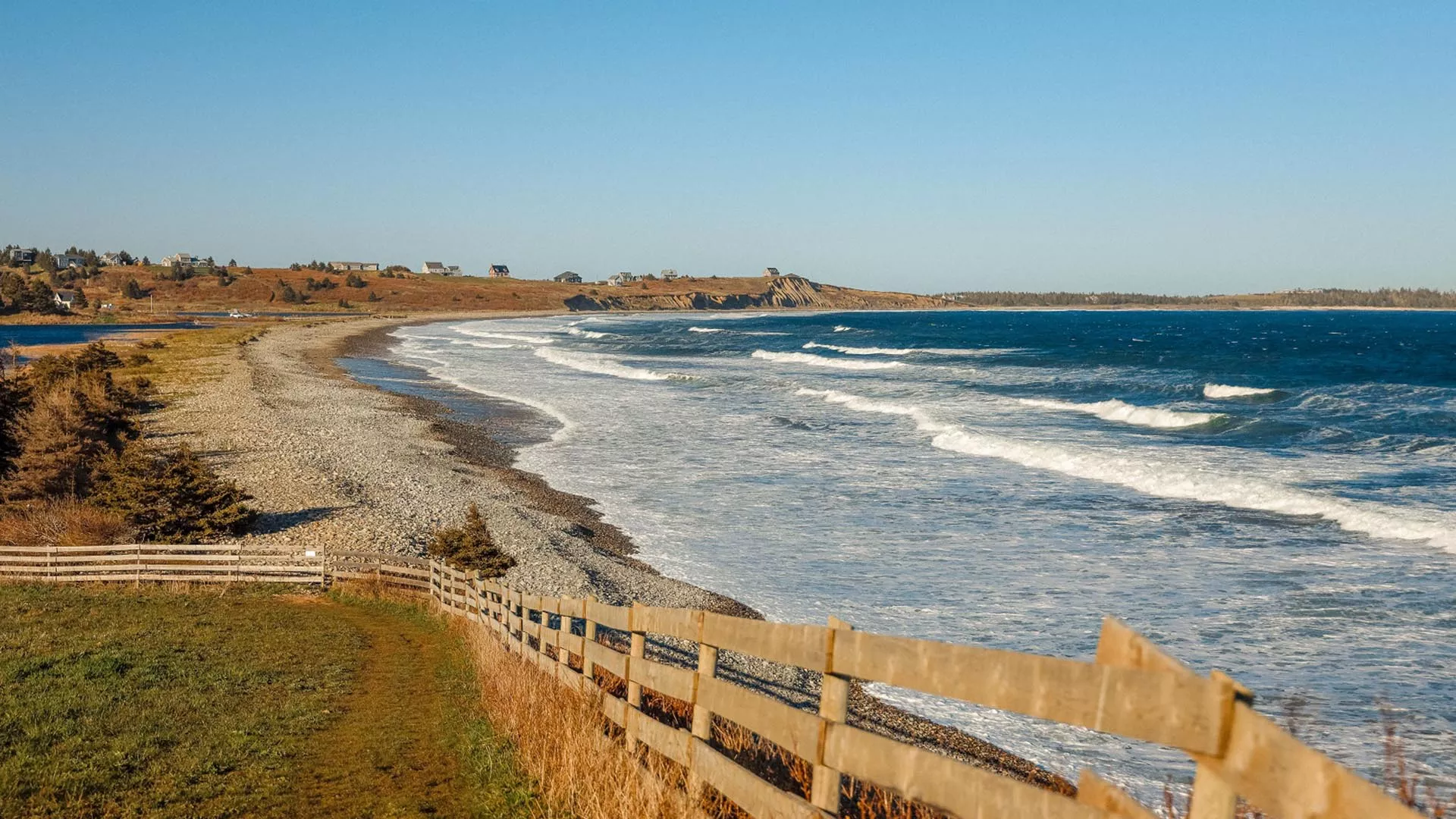 A scenic view of a pebbly beach with gentle waves, a grassy pathway with a wooden fence, and houses on a hill in the distance under a clear blue sky.