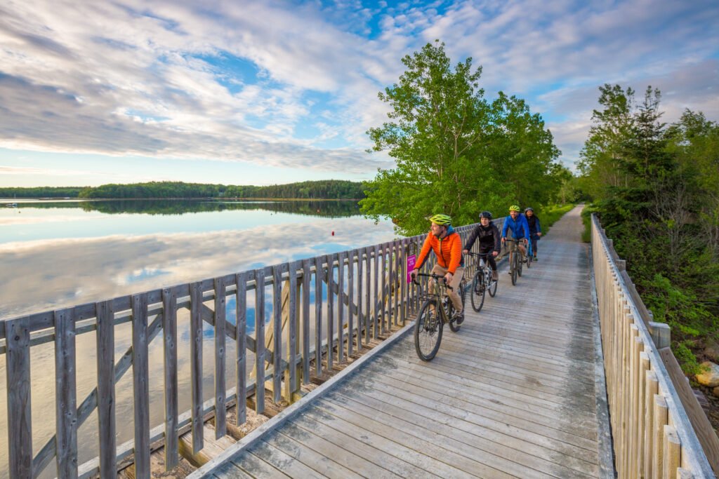 Four people riding mountain bikes on a wooden trail beside a calm lake with reflections of clouds and trees.