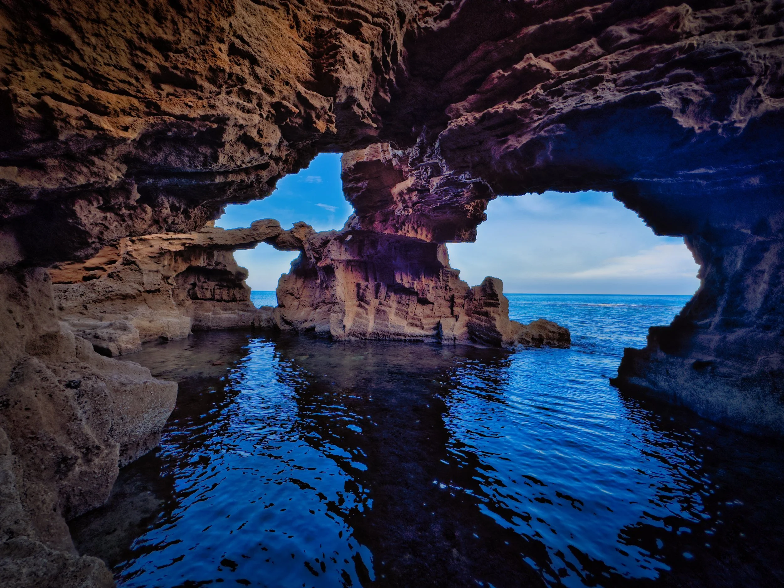 View of a rocky coastal cave with natural arch formations and blue ocean water, partly open sky visible through openings in the rocks.